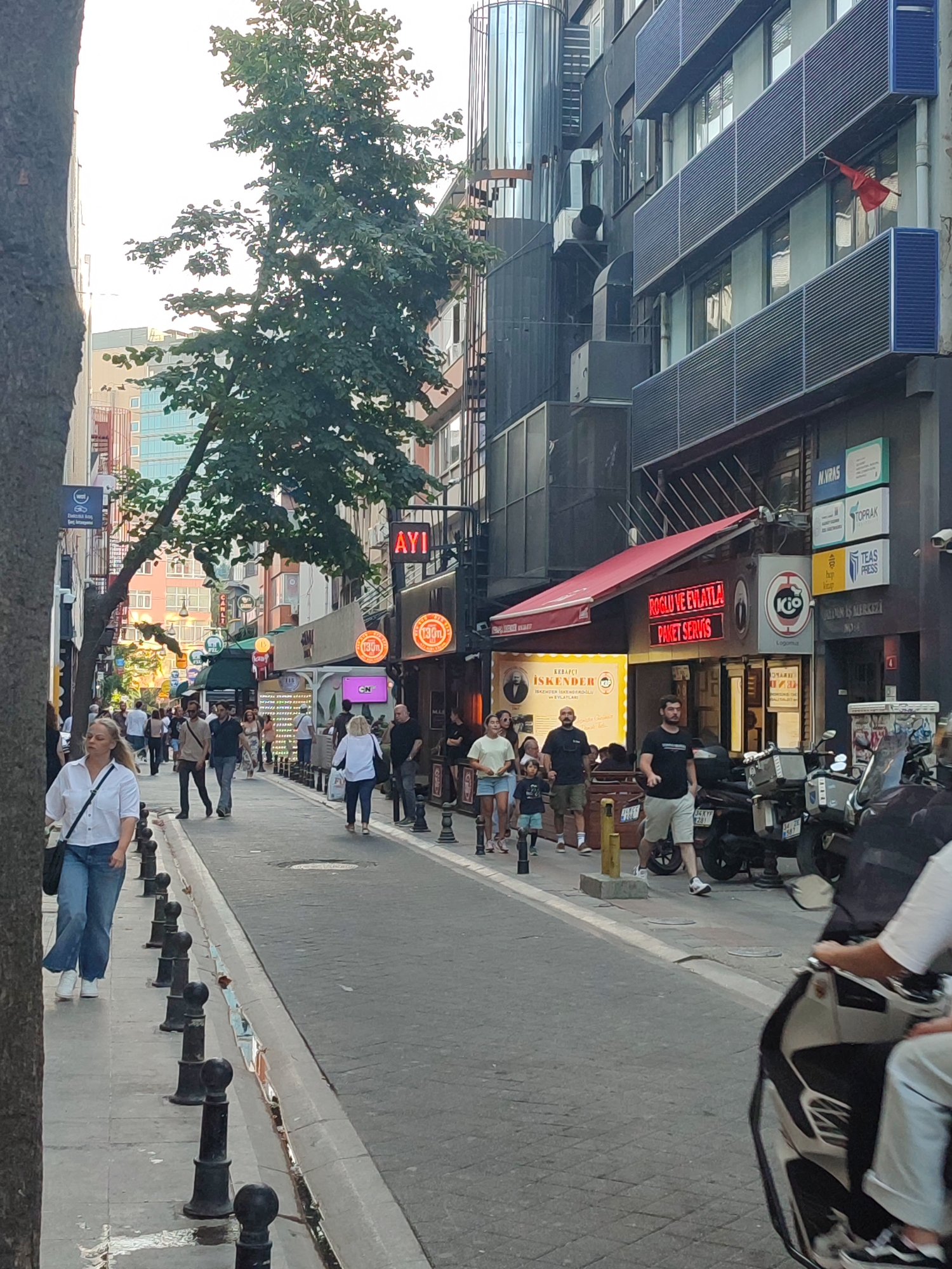 The image depicts a bustling street scene in Kadıköy, Turkey, likely during daylight hours. The street is paved with cobblestones, and lined with multi-story buildings on both sides, featuring a mix of architectural styles. Many buildings house shops and restaurants, indicated by visible signage, with awnings and outdoor seating areas. A prominent red neon sign reading "AYI" is visible.
Several people are walking along the sidewalk and crossing the street. They are dressed in casual attire, suggesting a warm day. Some individuals are walking alone, while others are in groups, including families with children. There are parked motorcycles and scooters on the side of the street, suggesting vehicle traffic.
A large tree with dense green foliage is growing on the left side of the street, partially obscuring the view of the buildings. The sky is overcast, contributing to the diffused lighting of the scene. The overall impression is that of a lively urban environment in a commercial district.