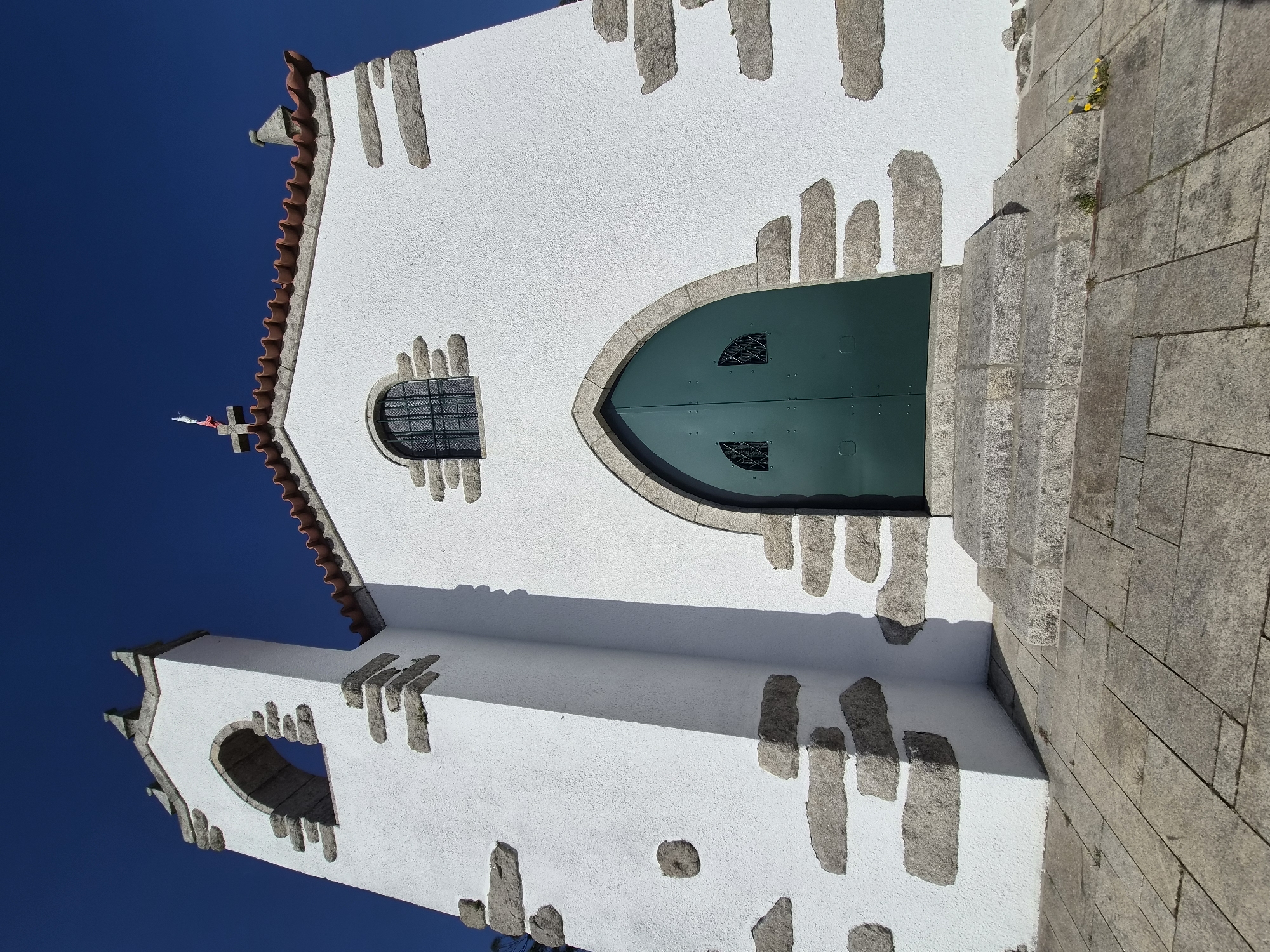 The image shows the side view of the Igreja da Misericórdia de Esposende, located in Esposende, Portugal. The church has a white exterior with gray stone accents. The front door is green and arched. There is a small, barred window to the left of the door. The roof is red tile. A bell tower is visible on the left side of the church. The sky is blue. The image is taken from a low angle and rotated counterclockwise. There are stone steps visible to the right of the door.