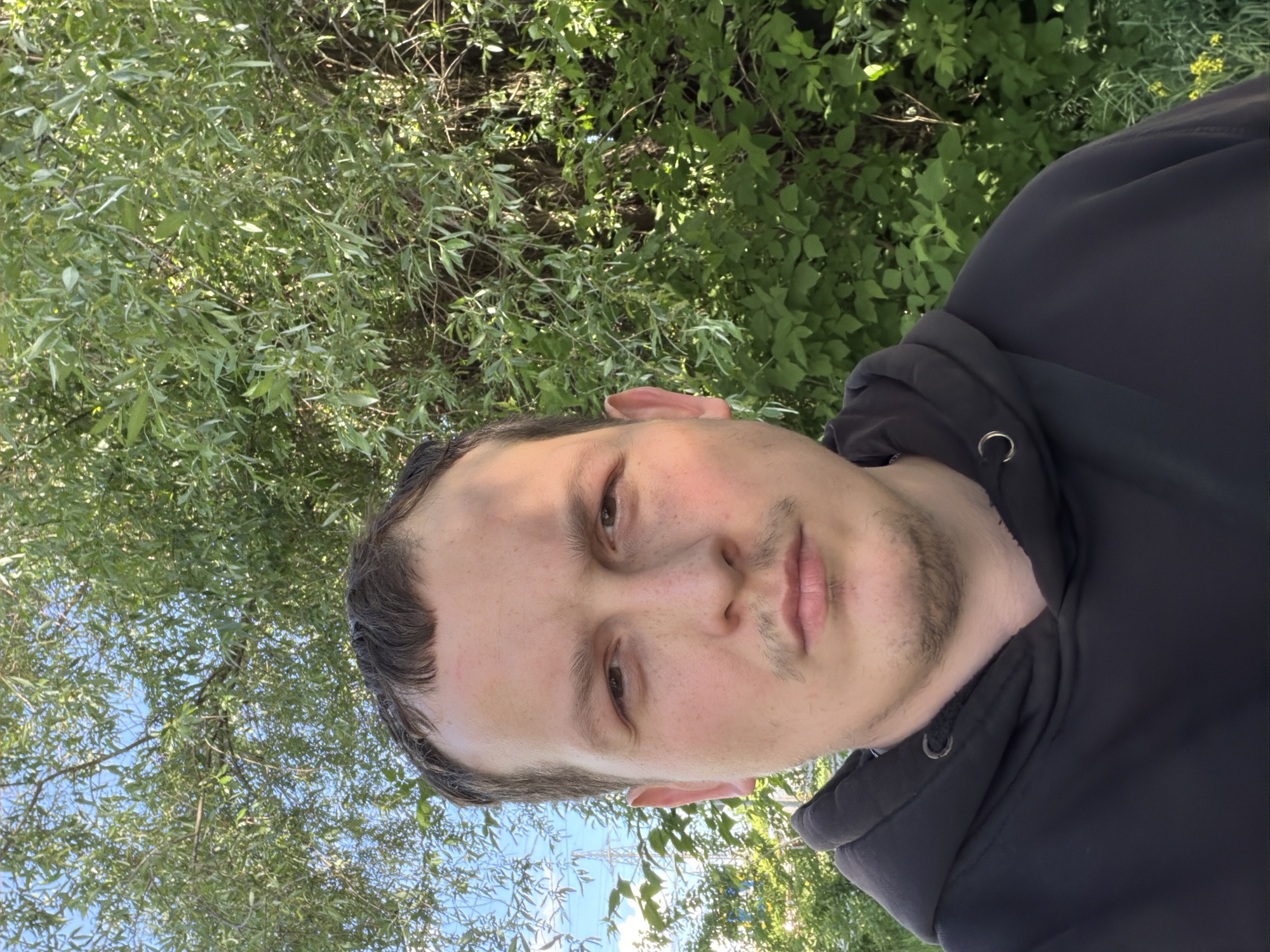 A man is posing for a selfie outdoors, partially obscured by foliage. He is wearing a dark hoodie. The background includes lush greenery, suggesting a park or wooded area. The location is Chaikovsky, Russia, as inferred from the surrounding clues. Electrical transmission towers are also visible in the background.