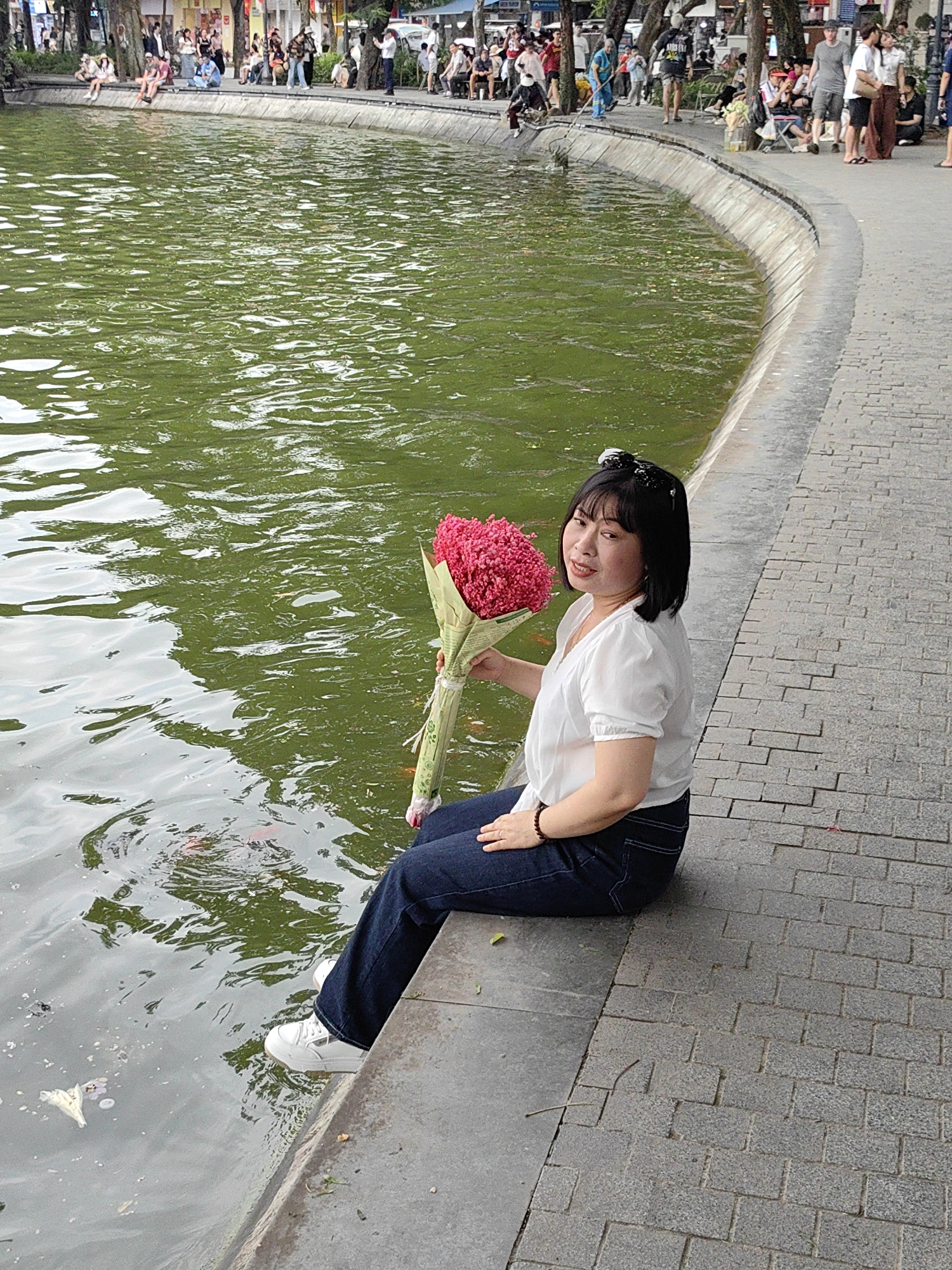 The image shows a woman sitting at the edge of Hoan Kiem Lake in Hanoi, Vietnam. The lake fills the left side of the image, with green water reflecting the sky. A woman is seated on the low concrete wall edging the lake. She wears dark jeans, a white short-sleeved blouse, and white sneakers. She holds a large bouquet of pink flowers wrapped in green paper. Her feet are close to the water's surface. The background contains a path, and behind the path, a crowd of people are visible, walking and standing. The day appears overcast.