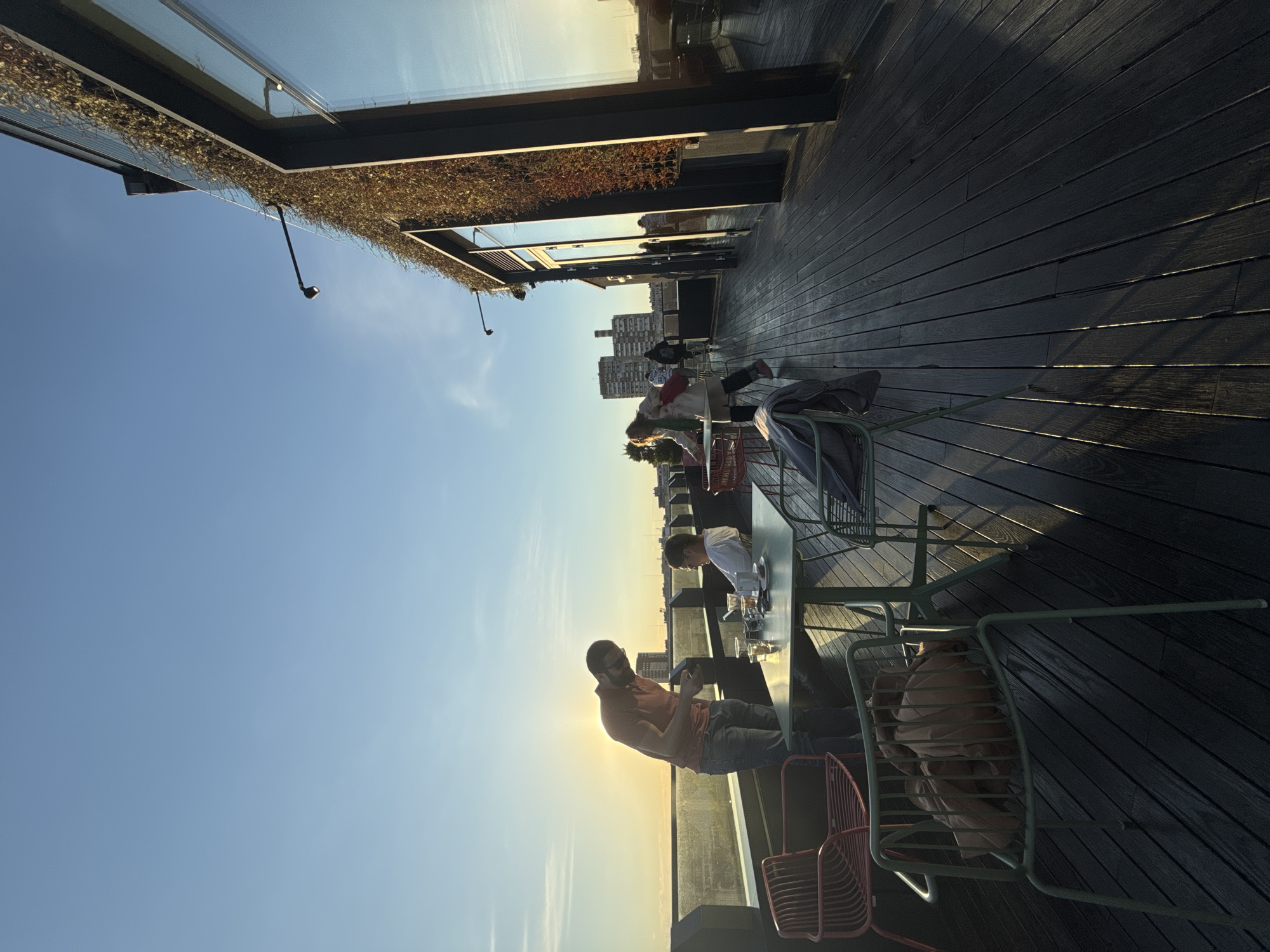A horizontal daytime shot depicts a rooftop bar setting in Sofia, Bulgaria. A black wooden deck forms the ground. At least four people are visible at different tables. One man, wearing an orange shirt and jeans, is seated at a table facing the setting sun, looking at his phone. At the next table, a man in a white shirt leans over the table, also appearing to be engaged with a phone or device. Behind these tables, other people are seated. In the background, mid-rise buildings characteristic of Sofia's urban landscape can be discerned. Above, a structure with a glass-topped roof, adorned with greenery, partially overhangs the scene. The clear blue sky transitions into a gradient of orange and yellow towards the horizon, indicating late afternoon or early evening light.