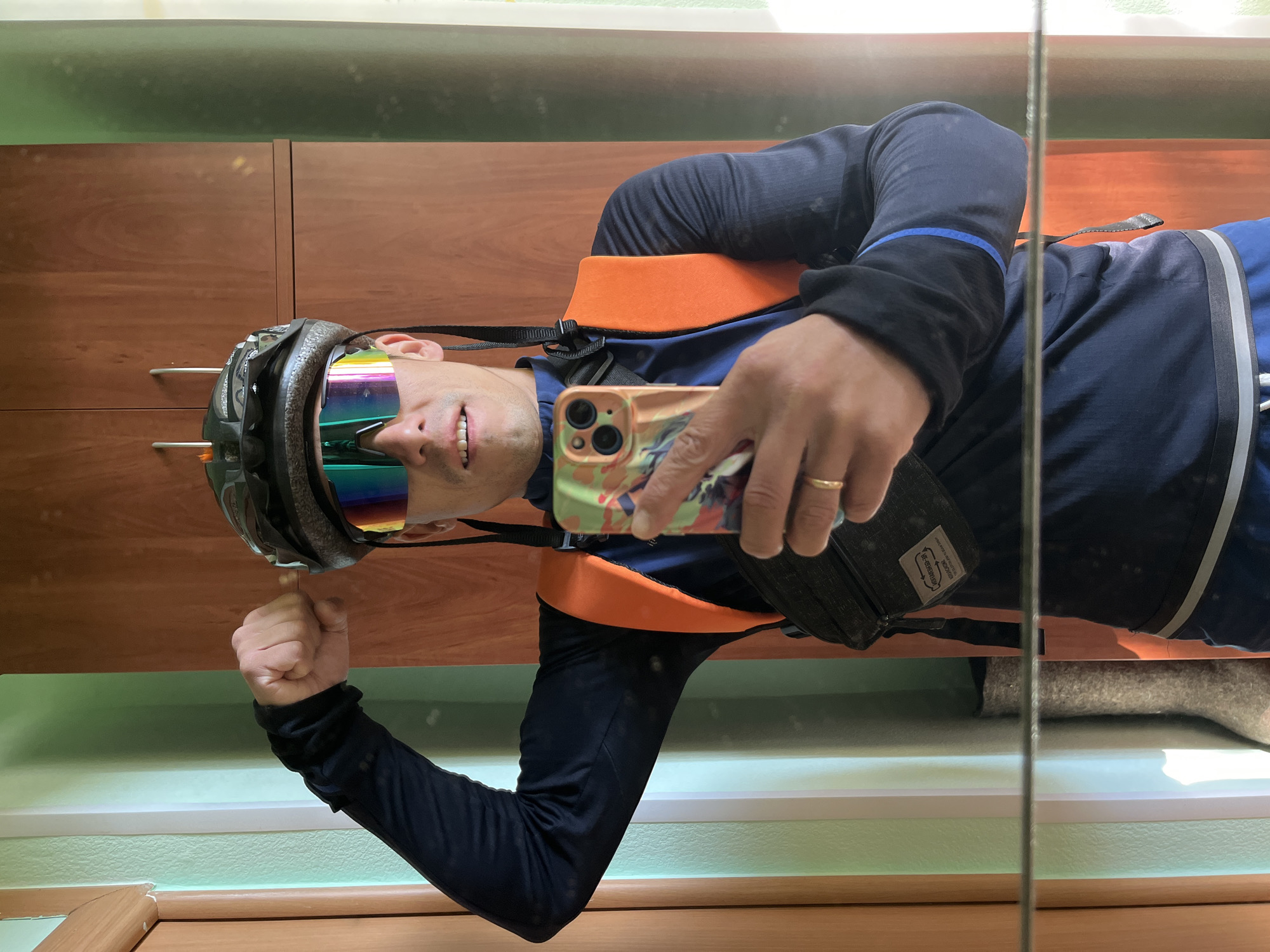 A man is taking a mirror selfie in what appears to be an apartment or house. He is wearing a dark blue long-sleeved shirt, blue shorts, a cycling helmet, and reflective sunglasses with a rainbow tint. He has an orange vest or backpack over his shoulders and a dark fanny pack or waist bag. He is flexing his left bicep and holding a smartphone in his right hand. The background shows a wooden door or cabinet and a wall with vertical green and white stripes. There is a reflection of a felt boot on the floor. The image does not contain any information or visual cues that would indicate its location in the city of Chaikovsky, Russia.