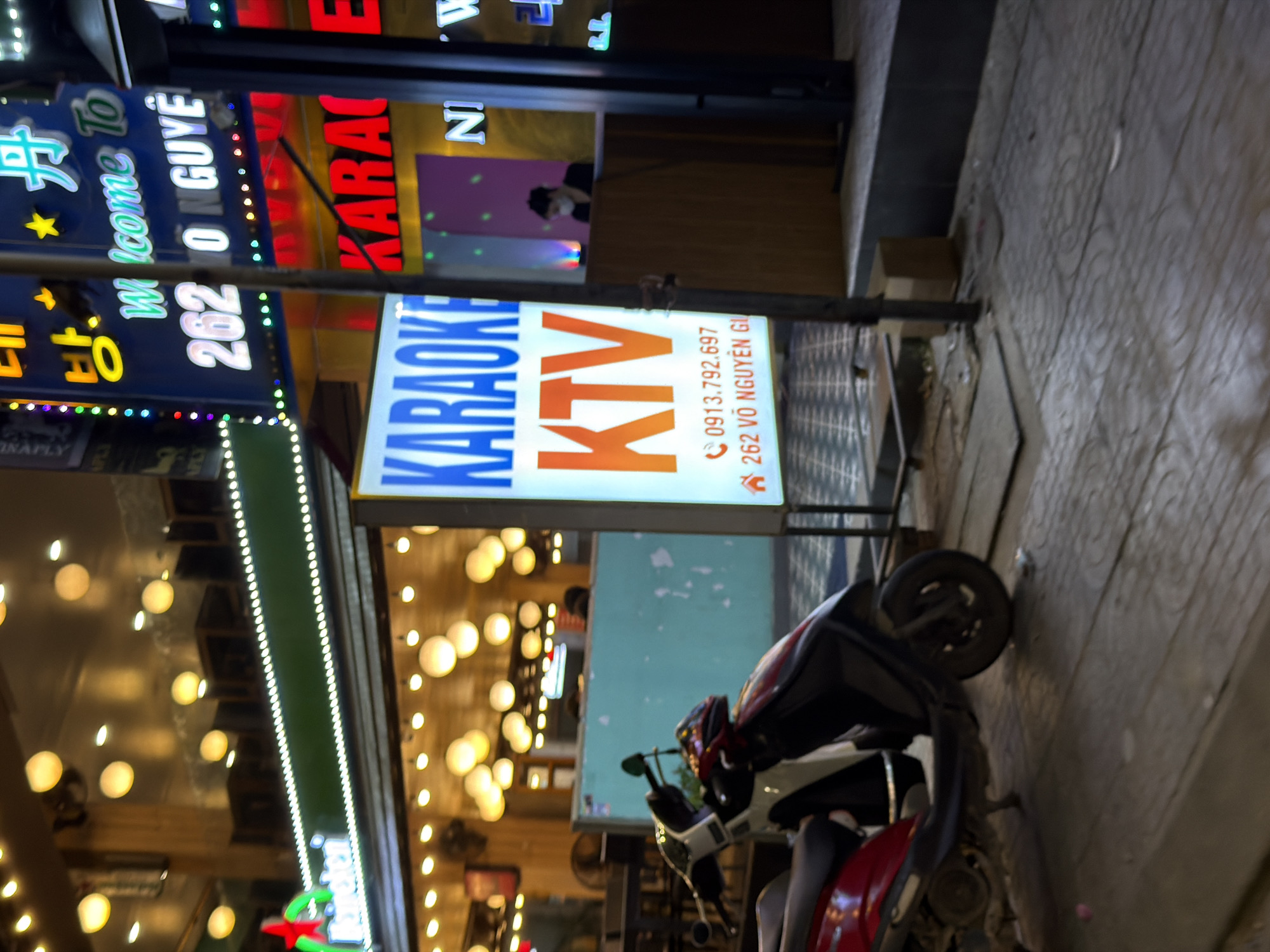 The image depicts a nighttime street scene in Đà Nẵng, Vietnam, likely in an area with entertainment venues. A red and white scooter is parked on a sidewalk paved with textured concrete. In the background, a brightly lit building features multiple signs advertising "Karaoke" and "KTV" in various colors, including neon blue and red. One prominent sign displays a phone number, "0913.792.697", and an address, "262 VÕ NGUYÊN GI". Above the Karaoke signs, a larger, more elaborate sign with Korean characters and the text "Welcome To" in green neon lights is visible. Further back, illuminated bulbs hang from the ceiling of the building's interior, suggesting a vibrant nightlife atmosphere. A person wearing a face mask and a dark top is visible behind a counter inside the establishment. The overall impression is one of a bustling entertainment district at night.