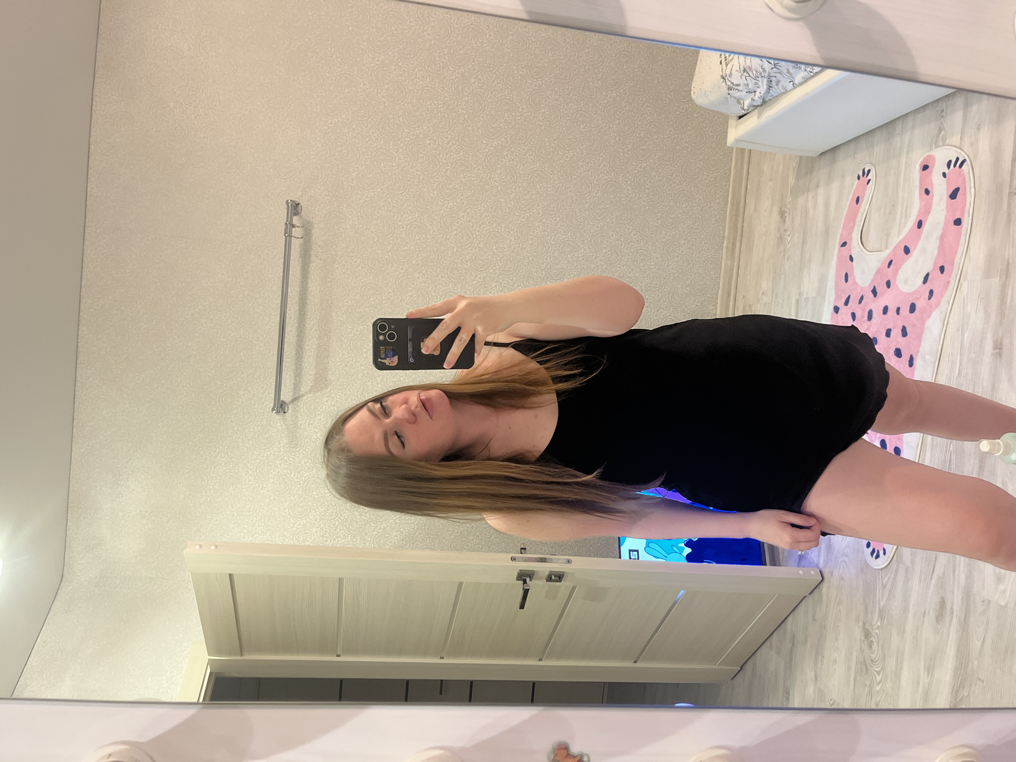 A woman stands in an indoor setting in Unknown, Unknown, holding a smartphone in her left hand and using it to take a selfie in front of a mirror. She wears a black dress. The room has textured wallpaper, a towel rack, a white door with a silver handle, and a light-colored wooden floor. A pink and black spotted rug lies on the floor. Behind the woman, part of a bed with white linens is visible.