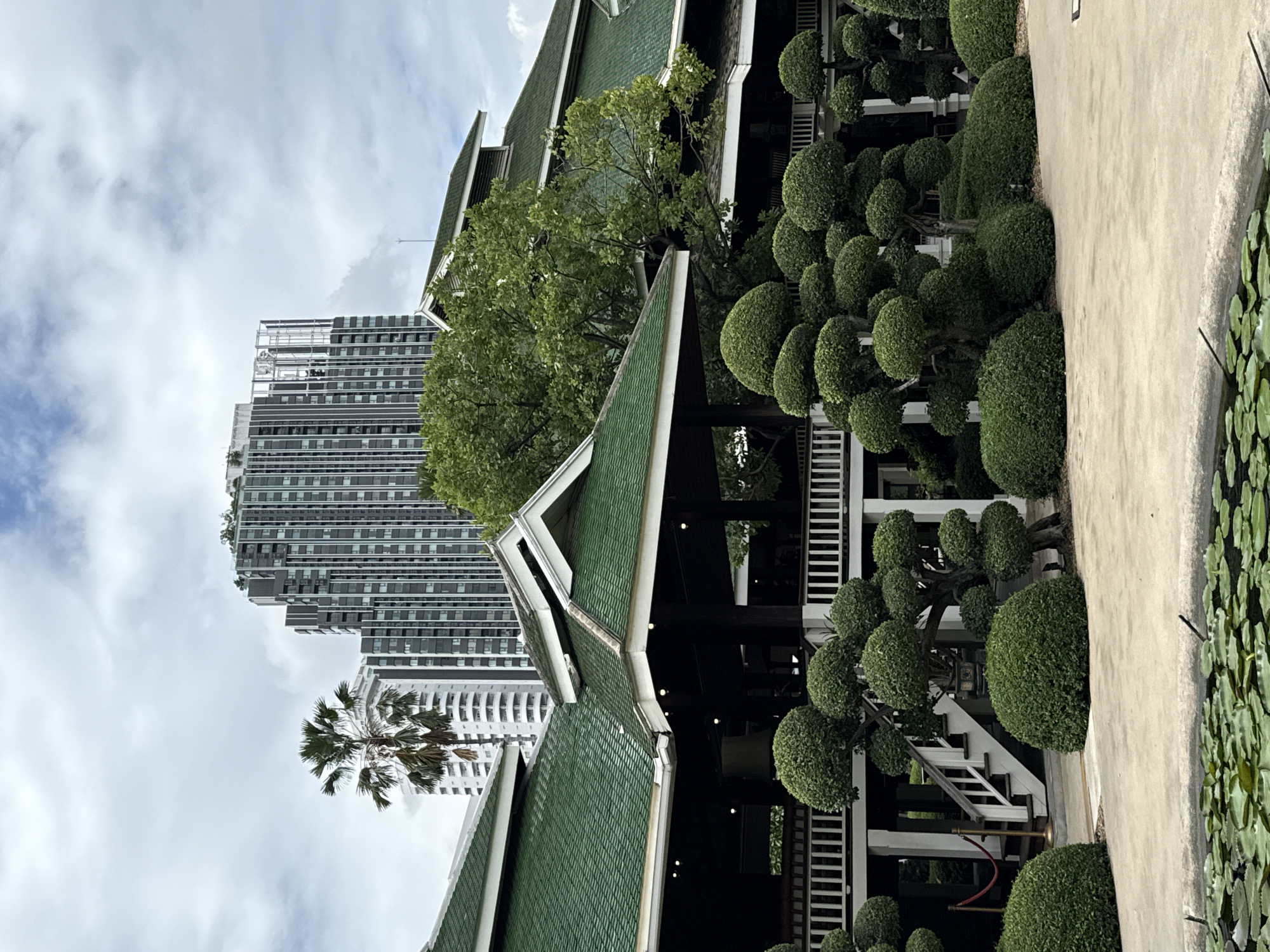 The image shows a traditional Thai-style building with a green tiled roof, contrasting with modern high-rise buildings in the background. The foreground features a garden with meticulously shaped topiaries and a pond with lily pads. A palm tree stands tall between the traditional architecture and the modern skyscrapers, suggesting a blend of old and new within the urban landscape of Bangkok, Thailand. The sky is cloudy, indicating an overcast day. There are no people visible in the image.