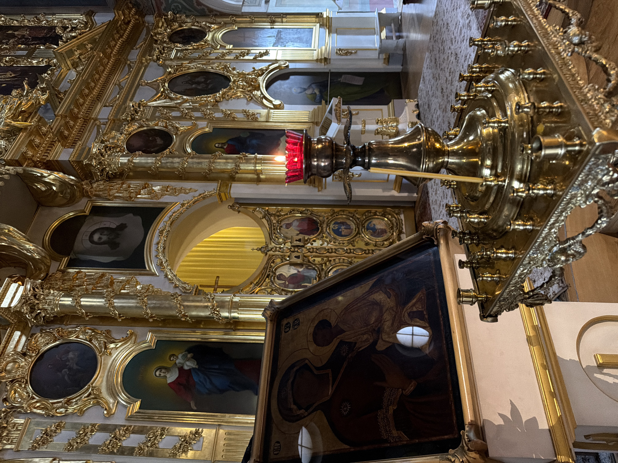 The image depicts the interior of a richly adorned Orthodox church, likely the Kazan Cathedral in Saint Petersburg, Russia, given the opulent gilded iconostasis and ornate decorations. In the foreground, a brass candelabra with a single lit red candle stands on a decorative brass base. To the left, an icon of the Virgin Mary and Christ Child is displayed prominently, leaning against a light-colored stand. The background reveals a section of the church's grand iconostasis, featuring numerous gilded frames around various religious icons and paintings. Yellow curtains are visible through an arched opening in the center of the iconostasis. The floor is covered with a patterned rug. The scene is characterized by its religious ambiance, intricate craftsmanship, and the warm glow of candlelight. There are no people visible in the image.