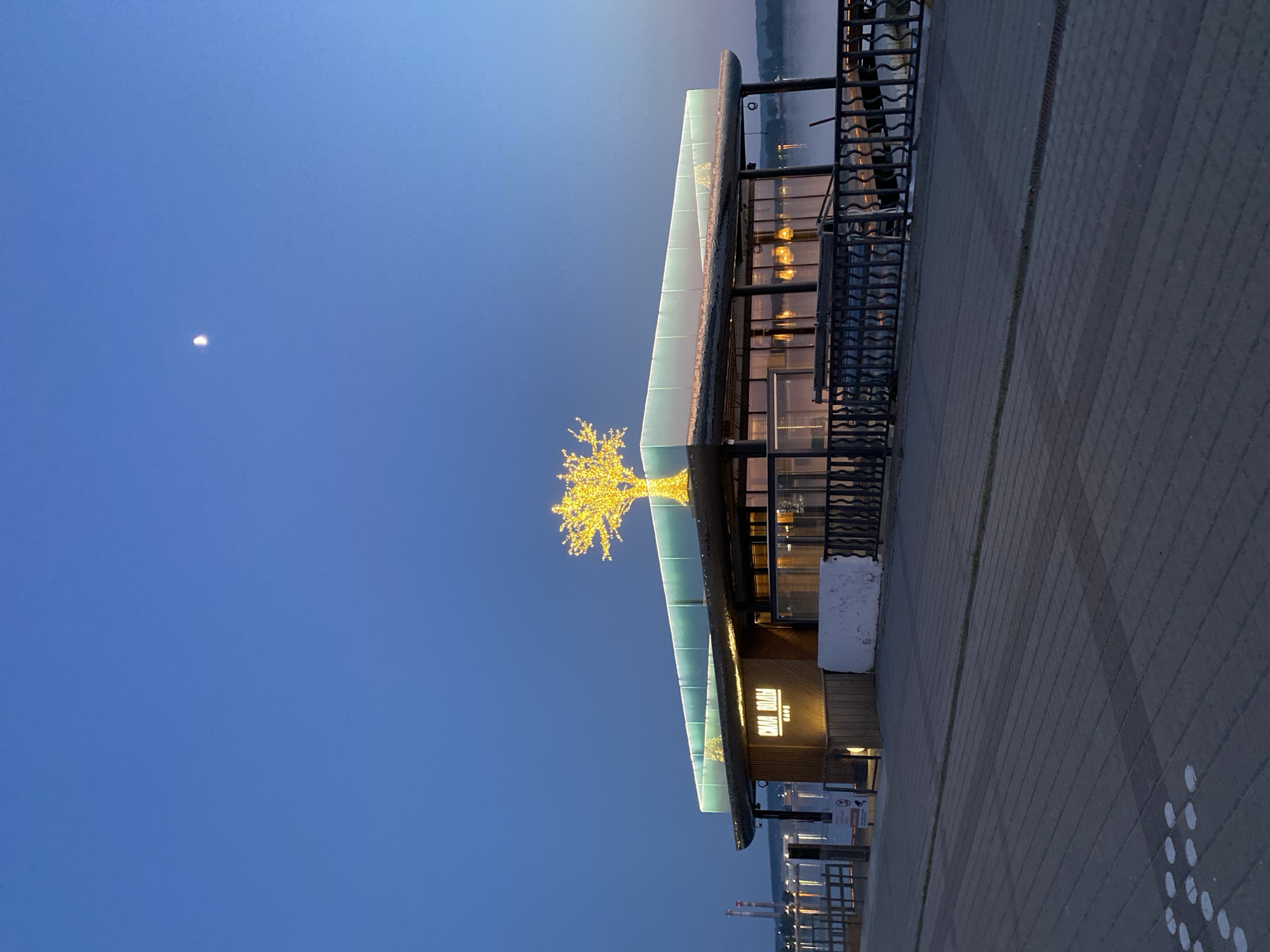 The image depicts a building at night in Votkinsk, Russia. The building is illuminated with warm, golden light, and a decorative tree made of lights is prominently displayed on its roof. The building appears to be a cafe or restaurant, with the sign "CHOCO ROOM" visible. The building is located on a paved walkway, and a body of water is visible in the background. The sky is a clear, dark blue, with a faint moon visible in the upper left corner. There is a metal railing along the side of the building. Some structural elements are seen in the background.