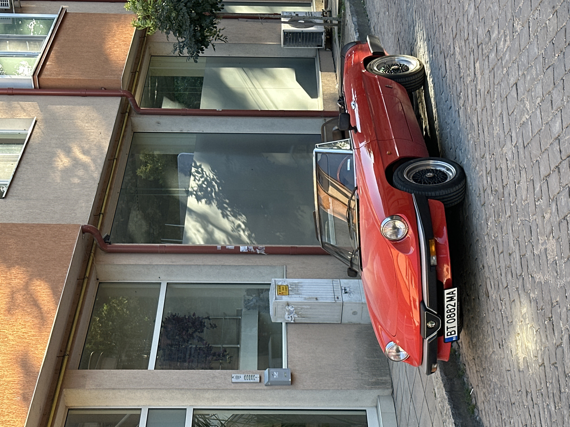 A bright red Alfa Romeo Spider convertible is parked on a cobblestone street in front of a building in Veliko Tarnovo, Bulgaria. The car, with its top down, has a license plate that reads "BT 0882 MA". The building behind the car has large windows and a textured, orange-toned facade. Shadows from tree branches are cast across the building's exterior and the street. An air conditioning unit is visible on the sidewalk to the right of the car. A red drainpipe runs down the side of the building. The overall scene suggests a sunny day in an urban environment.