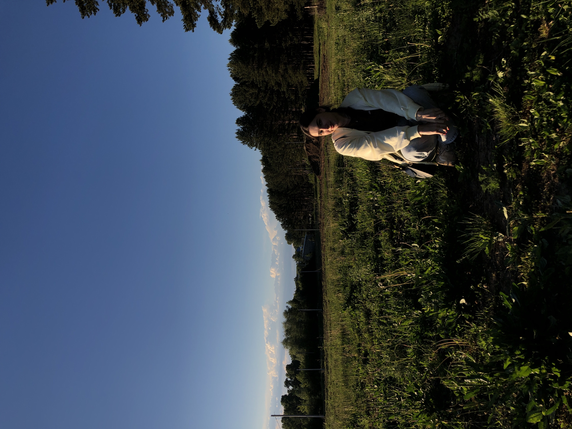 The image depicts a woman lying in a grassy field, possibly in a park. She is wearing light-colored clothing and is positioned approximately in the center of the frame. The background includes a forest line along the horizon and a clear blue sky.
While the image does not explicitly indicate its location in Chaikovsky, Russia, the combination of grassy terrain, a dense forest of coniferous trees, and a clear sky are elements consistent with the region's landscape. No immediate details confirm the exact location within Chaikovsky.