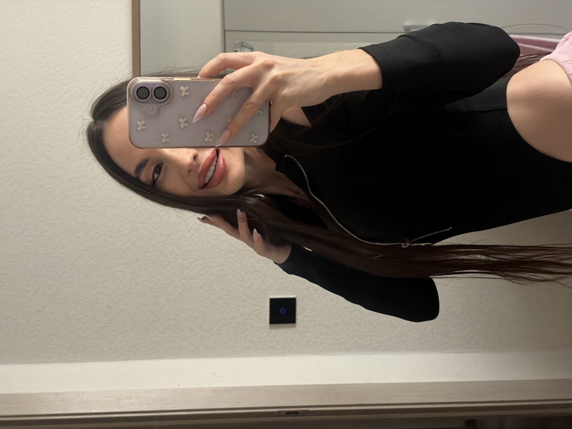 A person with long, dark hair is taking a selfie in front of a mirror. They are wearing a black top with a zipper. They have a light purple phone case decorated with white flowers. The background is a white wall with a dark square object, possibly a light switch. The location cannot be determined from the image.