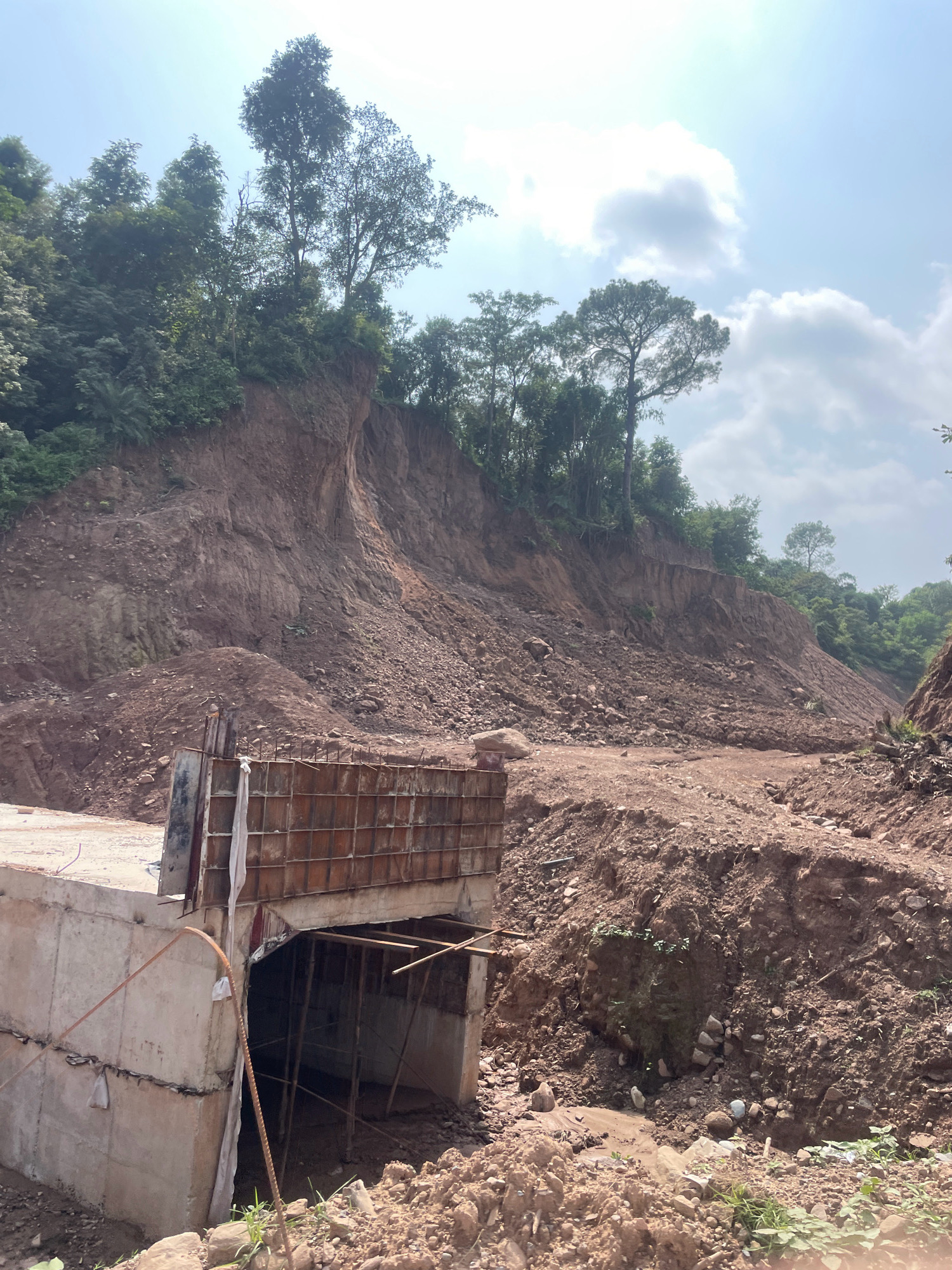 The image shows a construction site with a concrete structure in the foreground, likely a bridge or culvert under construction. The structure is made of concrete and reinforced with rebar. Behind the structure, a steep, eroded hillside covered in loose soil and rocks dominates the scene. Trees and vegetation grow along the top and sides of the hillside. The sky is bright and partly cloudy. The scene suggests ongoing construction work in a natural, hilly environment. The location is identified as Jaanta, India.
