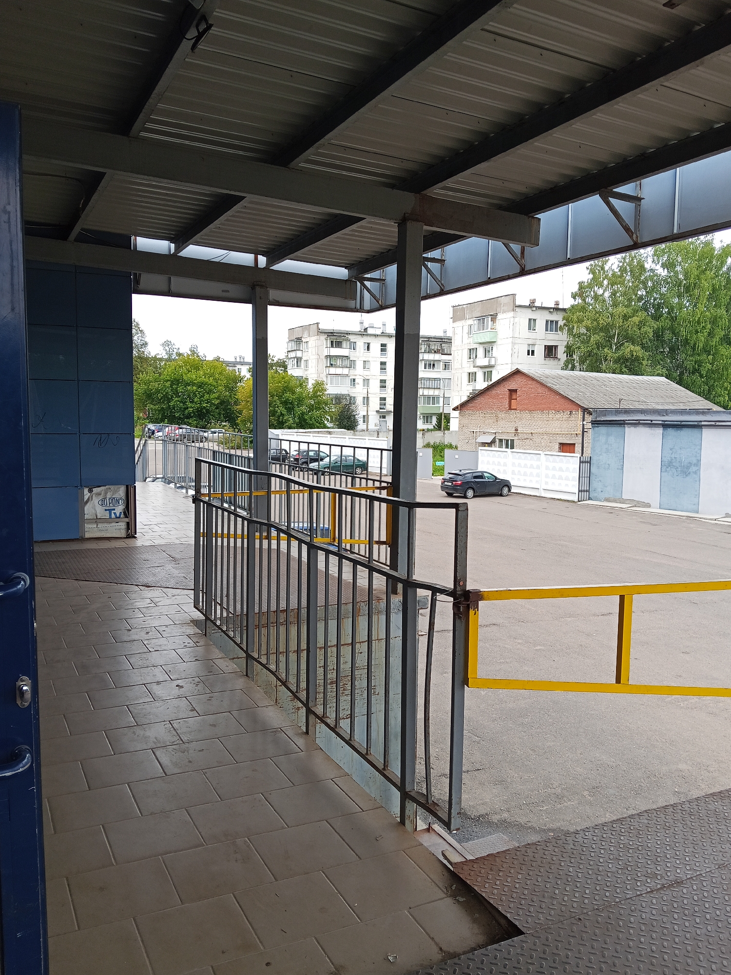 The image shows a covered walkway with a tiled floor leading to an outdoor area with a metal fence and a yellow barrier. In the background, there are several apartment buildings and a smaller brick building with a gray roof. Trees are visible between the buildings. Cars are parked in the outdoor area. The scene appears to be a public space, possibly a bus stop or an entrance to a building complex. The location is identified as being in the city of Chaikovsky, Russia.