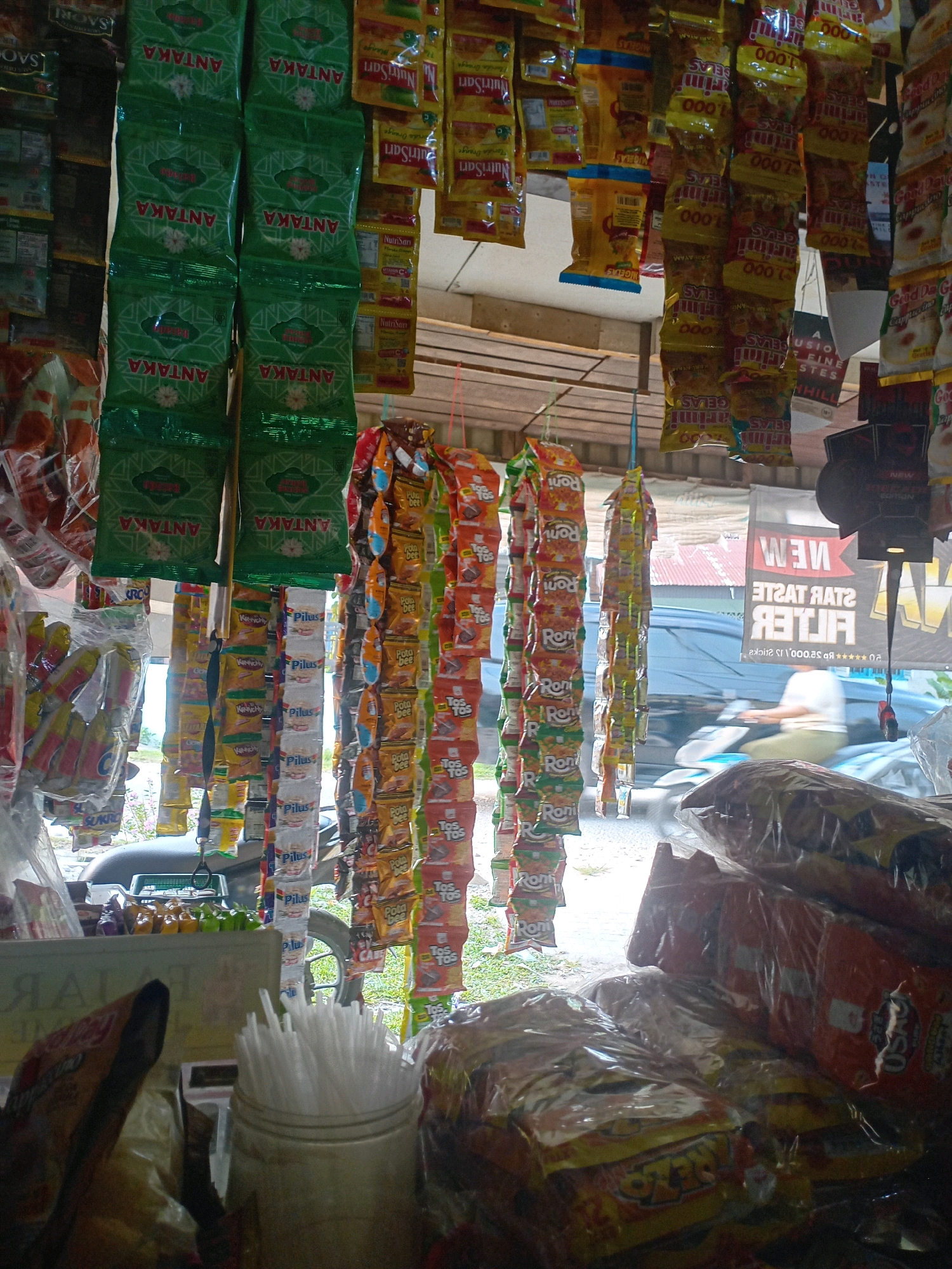 This is a low-angle, medium shot of a small street-side shop in Pekanbaru, Indonesia. The shop appears to be packed with merchandise, primarily snack food items. In the foreground, there are several large plastic-wrapped bags of snacks piled up, some with orange and yellow packaging featuring text in what appears to be Indonesian. To the left, there's a close-up of a collection of individually wrapped items, some in green packaging with the word "ANTAKA" visible. To the right and center, numerous small, colorful snack packets are hanging from strings, creating vertical rows. These packets display various brand names and colorful graphics. In the mid-ground, through an opening, a street scene is visible with a vehicle, possibly a scooter, passing by. The overall impression is of a bustling, local convenience store with a wide variety of affordable goods.