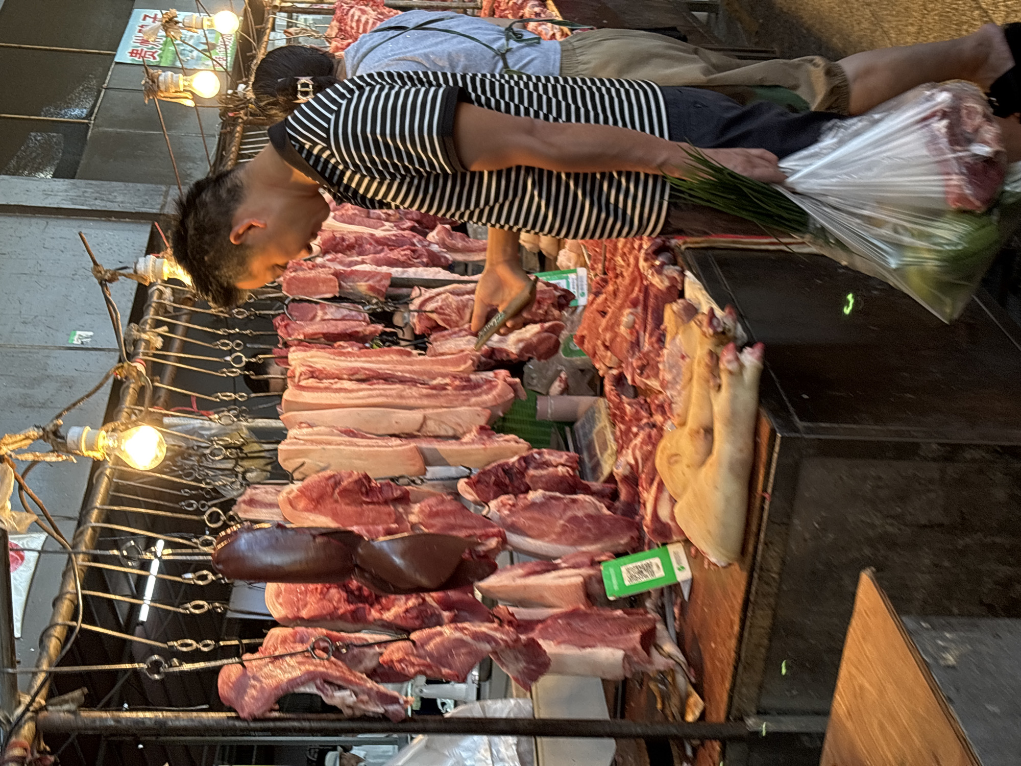 A man in a striped shirt holds a plastic bag containing green onions and meat while standing at a meat market. He is looking at his phone, which he is holding in his right hand. A QR code is visible on a green card on the counter. The man is facing away from the camera. Behind him, a woman wearing a grey shirt and an apron is visible. The market appears to be outdoors, with meat hanging from hooks overhead. This image was taken in the city of Jiangjin, China.