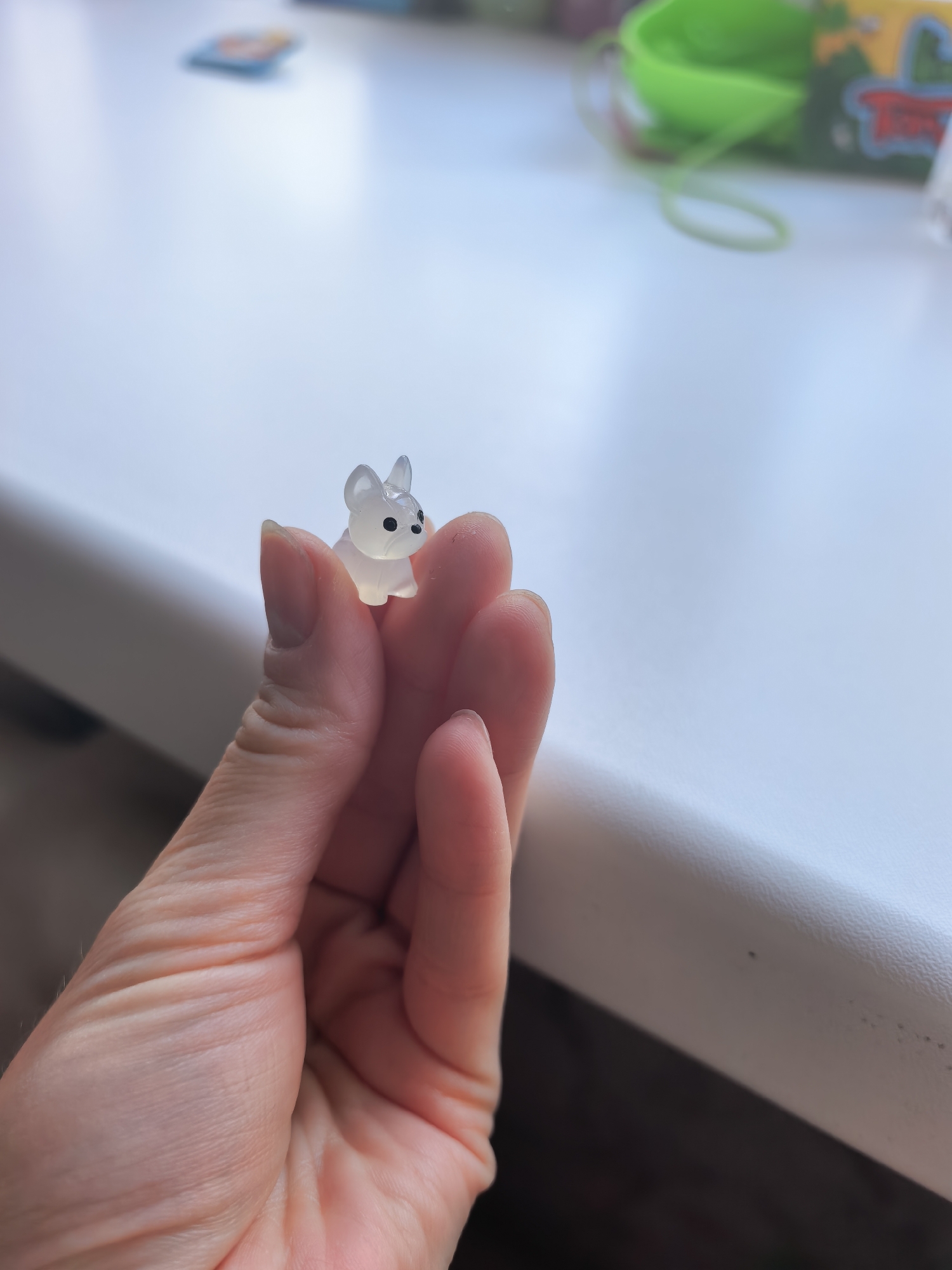 A hand holds a translucent, miniature dog figurine with black eyes. The background is a white surface, possibly a windowsill, with some blurred objects in the distance. The location is not identifiable as Sterlitamak, Russia, from the visual information provided.