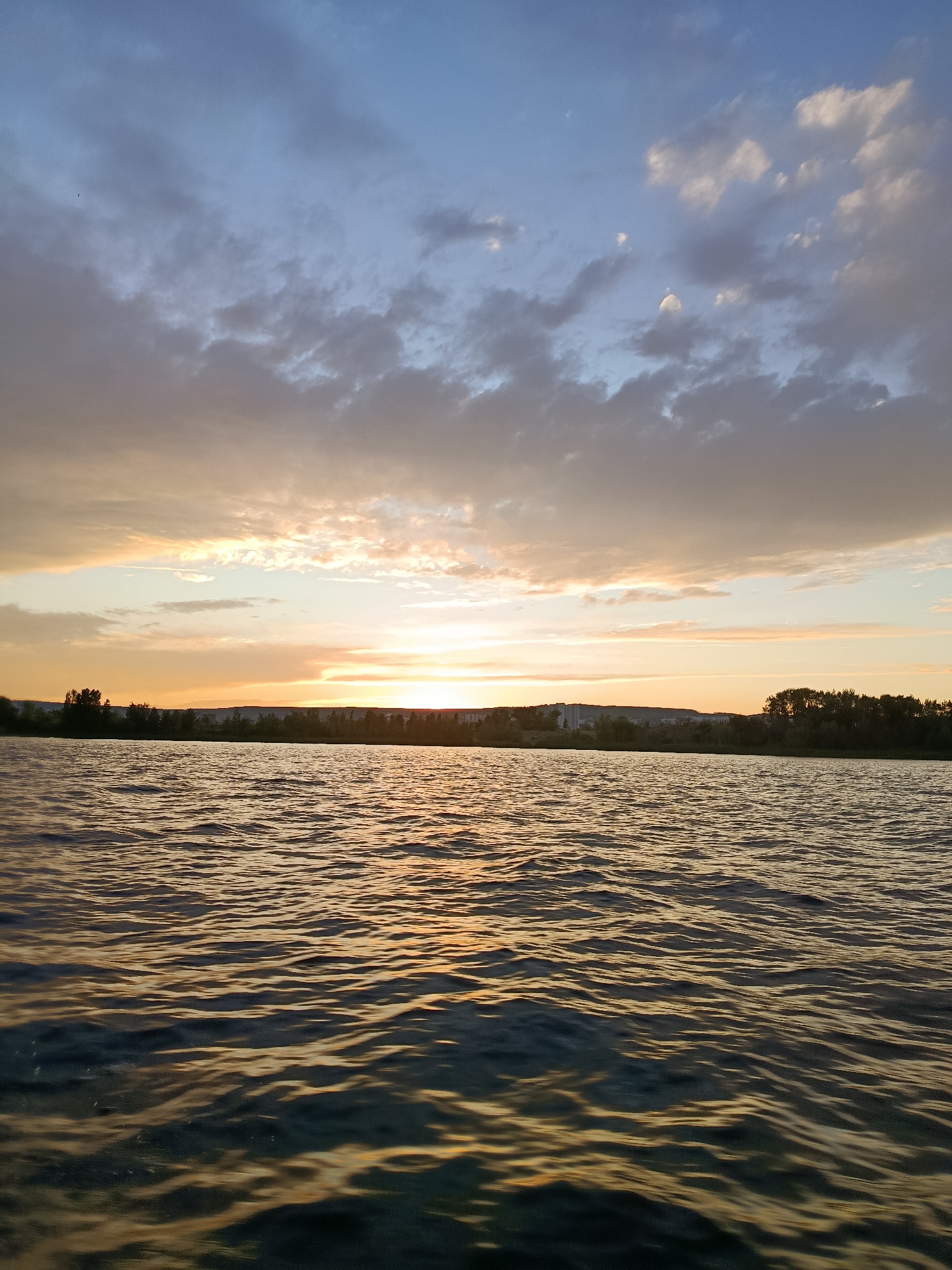A wide shot captures a body of water during sunset. The sky is a blend of soft blues and oranges, with scattered clouds that are illuminated by the setting sun. A thin line of trees and buildings marks the distant horizon. The water's surface is rippled, reflecting the warm colors of the sky, creating a shimmering effect. The overall scene is serene and depicts a natural landscape. This scene is located on the Volga River in Saratov, Russia.