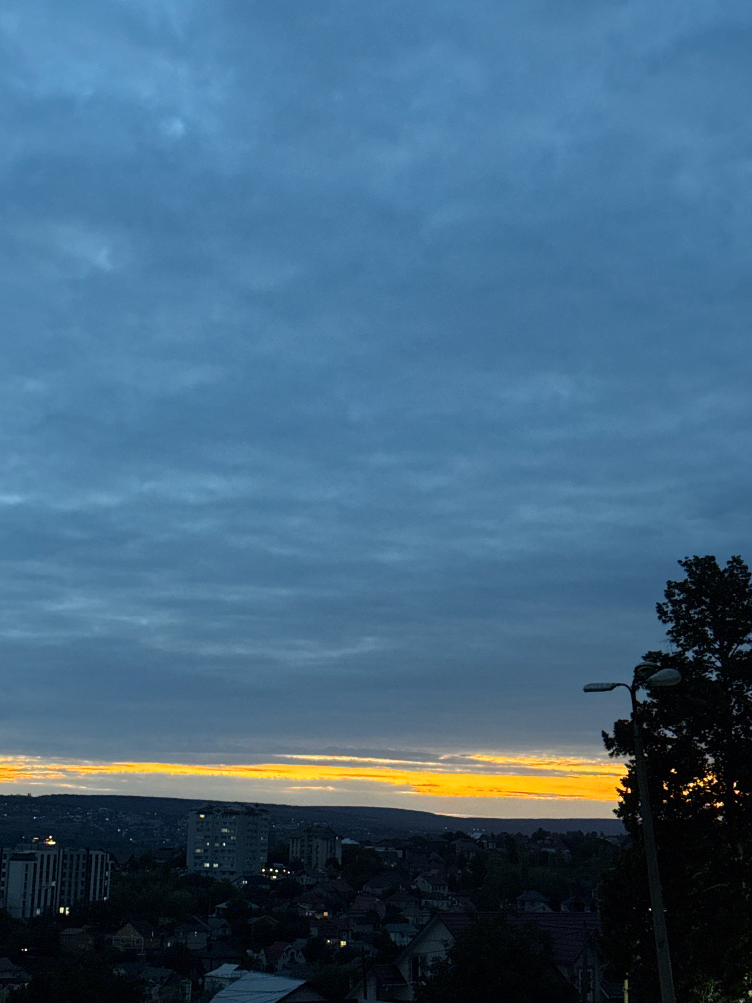 An exterior, medium shot captures a panoramic view of the city of Ialoveni, Moldova, under a moody, overcast sky just before sunset. The sky is a deep, textured blue, with horizontal cloud formations suggesting an approaching dusk. A striking band of vibrant orange and yellow light cuts across the horizon, indicating the low position of the sun.
In the foreground, a silhouetted tree dominates the right side of the frame, its dark foliage obscuring the sky. A street lamp stands to the left of the tree, also in silhouette, with a white lamp head visible.
Below the sky, the city of Ialoveni unfolds. The cityscape is a mix of residential buildings, with some multi-story structures displaying lit windows, and smaller houses. The overall impression is of a town winding down for the evening. The terrain appears slightly hilly, with the city nestled within the landscape. The image is a still, quiet scene, devoid of human activity or visible movement, emphasizing the natural light and the urban environment.