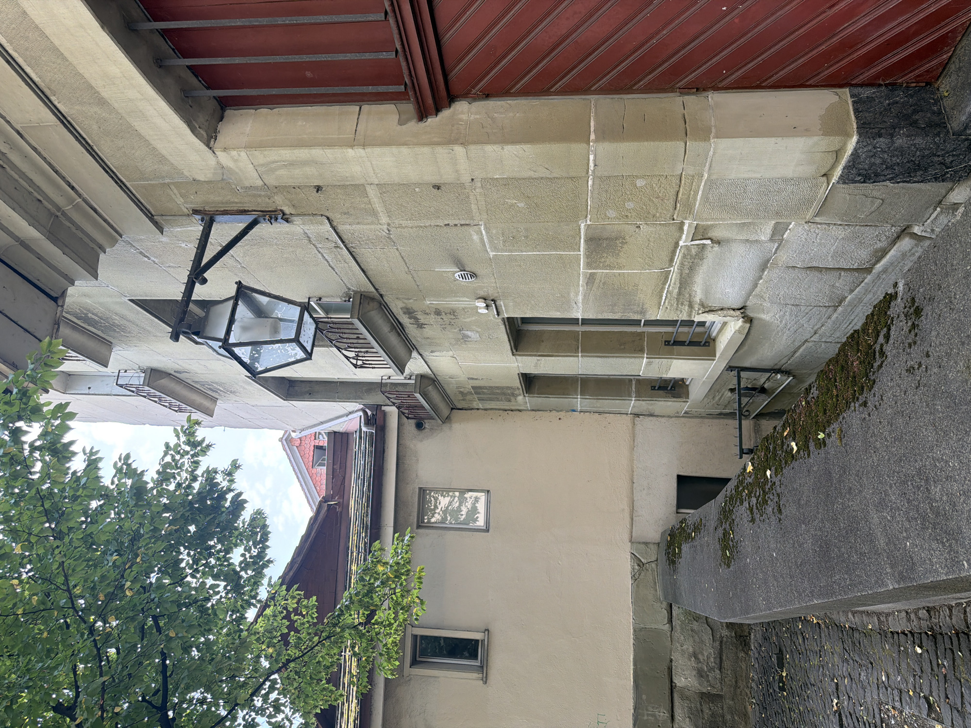 The image captures a narrow cobblestone street in Bern, Switzerland, bordered by a stone wall. A large, ornate lantern with a hexagonal glass body is mounted on the exterior wall of a building, which is constructed of large, light-colored stone blocks. A red double door with vertical planks is visible on the right side of the frame. The building has several windows, some with metal gratings, and small balconies with railings. Trees with green leaves overhang the scene from the left. The sky is cloudy.