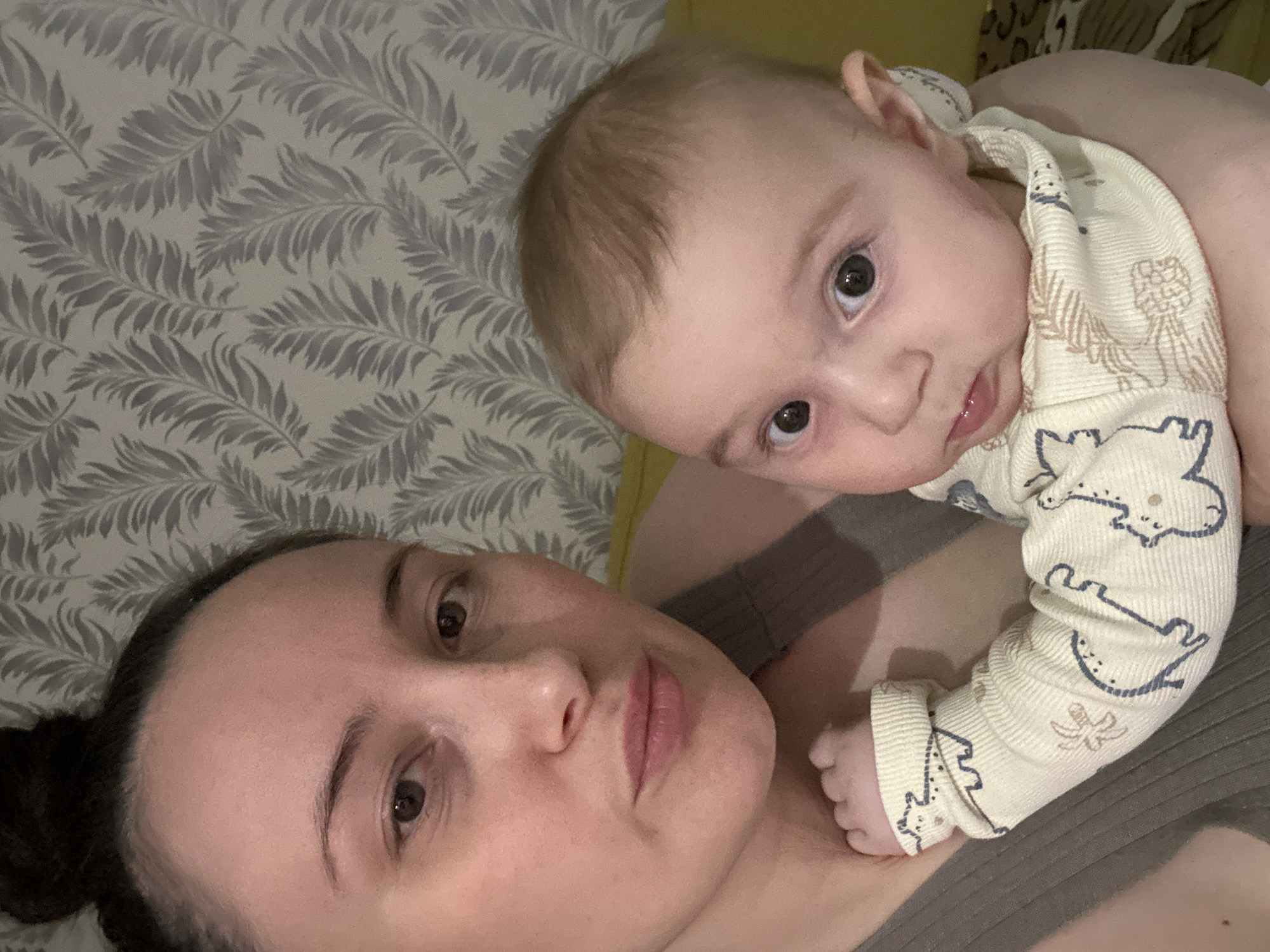 A woman with dark hair pulled back in a bun is lying down, presumably on her back, next to a baby. The baby is positioned on her chest and is wearing a light-colored romper with a dinosaur and banana pattern. Both the woman and baby are looking at the camera. The background includes a wall with a gray and white leaf pattern. The location is Chaikovsky, Russia.