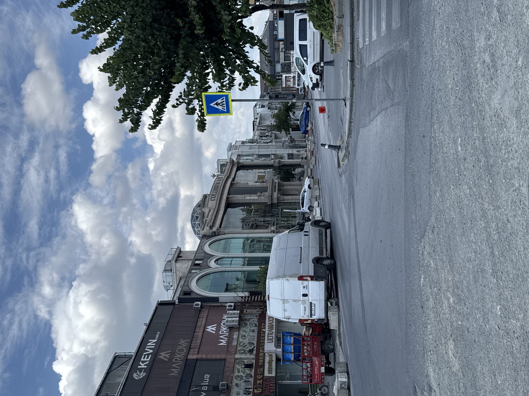 This is a street-level view in Gungoren, Turkey, on a cloudy day. The scene is dominated by a paved road with a pedestrian crossing marked by white stripes. In the foreground, the road surface appears somewhat worn, with patches of gravel and tire marks.
On the left side of the road, a white van is parked, its rear facing the camera. Adjacent to the van is a small red three-wheeled utility vehicle loaded with blue containers. Further back on the left, several commercial buildings line the street, displaying various signs including "KEVIN.com.tr", "MARKOVA", and "ULTRAMAXX". These buildings have modern architectural elements with large windows and some metallic cladding.
On the right side of the road, a pedestrian crossing sign with a yellow border and a blue center depicting a walking figure is mounted on a pole. A lush green tree with dense foliage is positioned to the right of the sign, partially obscuring the background. Further down the street, more buildings are visible, including structures with arched windows and ornate facades, one of which has "COLLISIUM MALL" signage. Several white cars are parked along the street, and a blue car is seen in the distance. The overall environment suggests an urban commercial district.