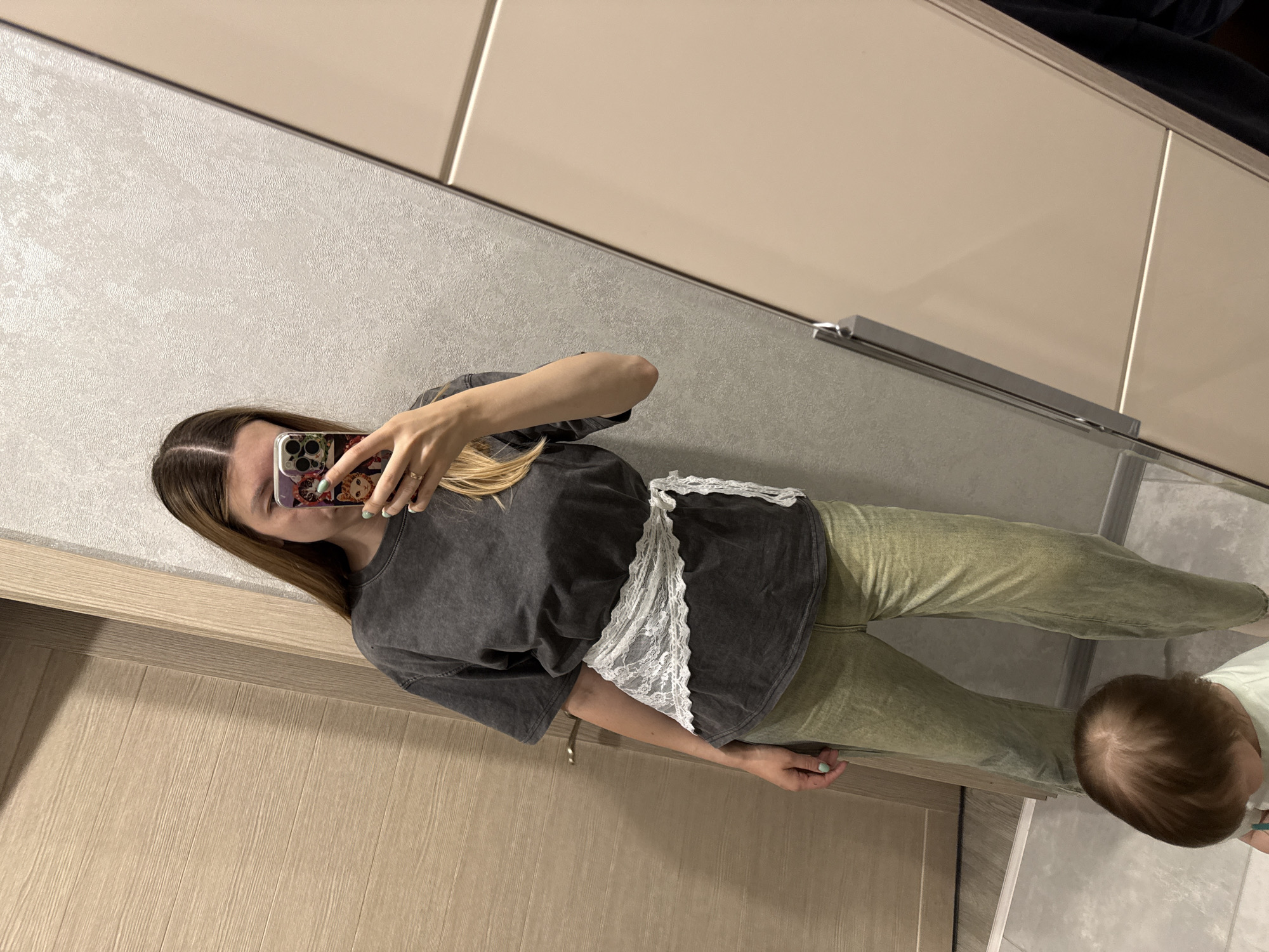 A woman is taking a selfie in what appears to be a bathroom or closet setting. The woman has long brown hair and is wearing a dark gray top with white lace detail and green pants. She holds her phone up, partially obscuring her face. In the lower right corner of the image, there is a baby with brown hair. The environment includes a light-colored door and what might be cabinets. The location is not identifiable as Moscow, Russia, without further information.