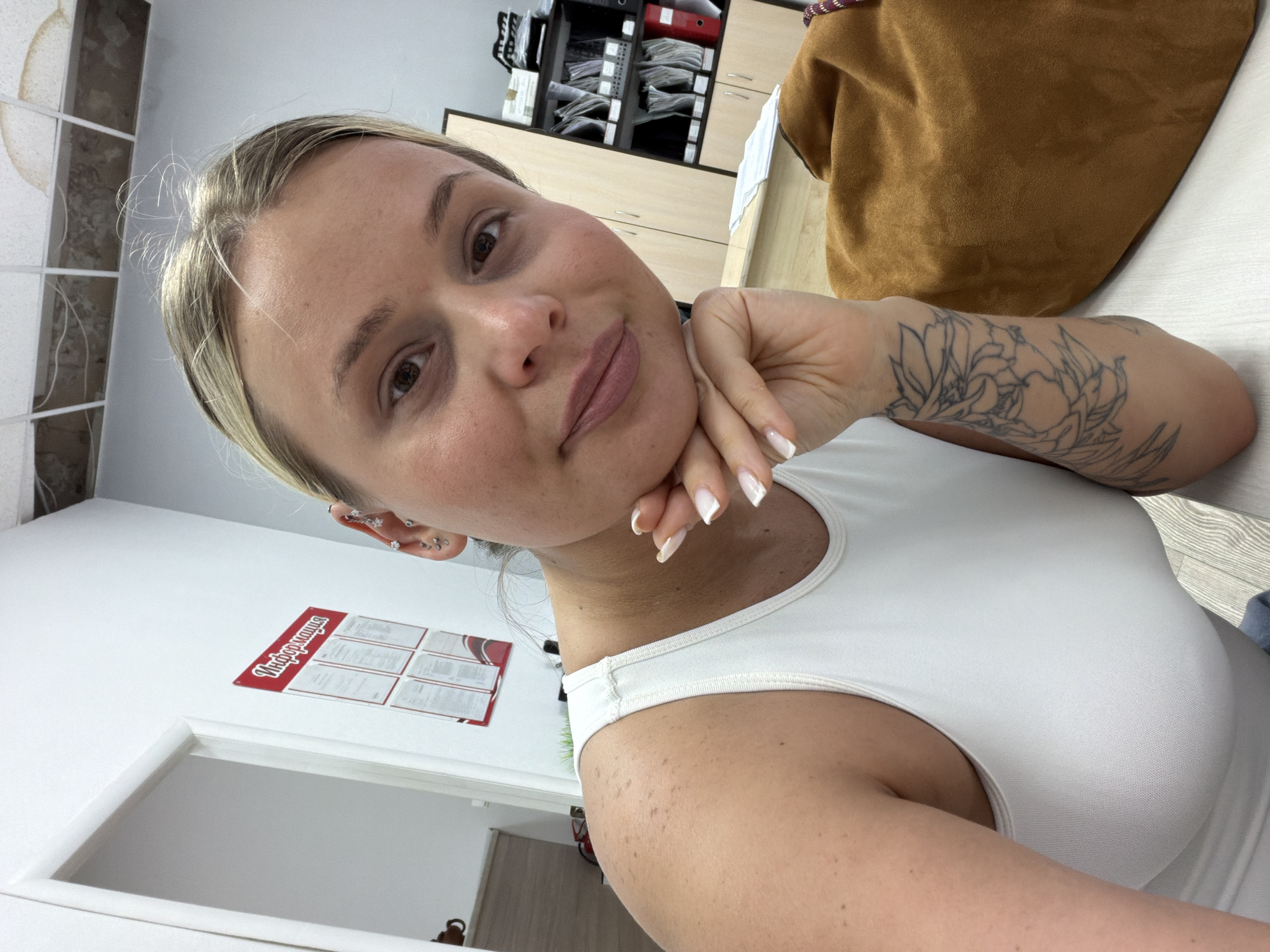 A woman with blonde hair and brown eyes poses for a selfie. She wears a white tank top. Her left arm is visible, featuring a floral tattoo. She rests her chin on her left hand. Her fingernails are manicured. She has multiple earrings in her left ear.
The background includes office-like elements: filing cabinets with folders and papers, a beige coat or bag, and an informational sign in Russian. The location is likely an office in Sarapul, Russia, based on the signage in the background. There is a doorway in the background as well.