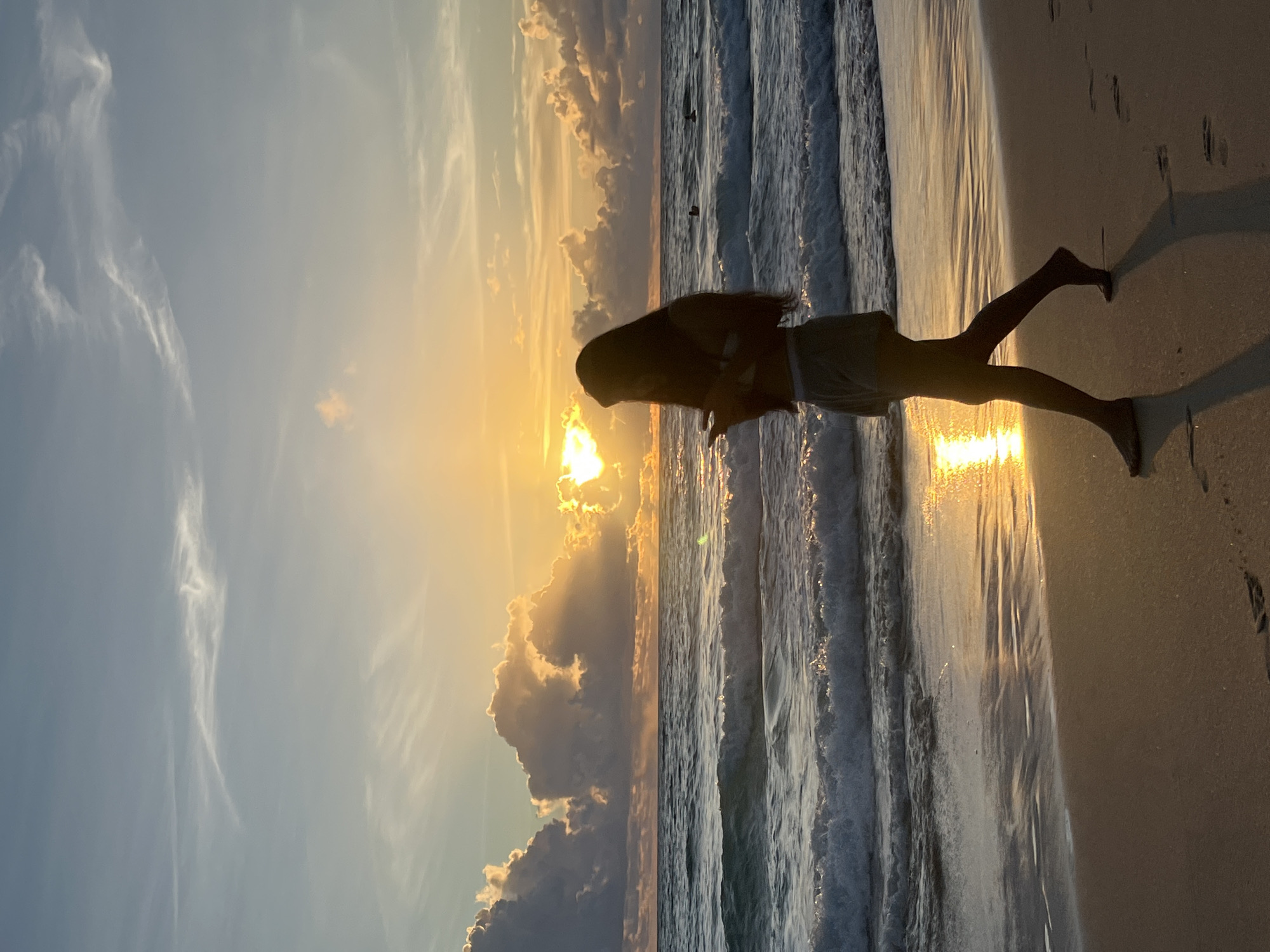 A full-body, silhouetted shot shows a person walking along a wet sandy beach. The person is facing left and is in the middle of a stride, with their phone in their hands. They are wearing shorts and a tank top. The ocean is to the left of the person, with waves breaking on the shore. The sun is setting or rising on the horizon, casting a golden glow on the water and clouds. The sky is a gradient of blue and orange, with scattered clouds. The beach is sandy and wet, with footprints visible in the sand. The location is a beach in Choeng Thale, Thailand.
