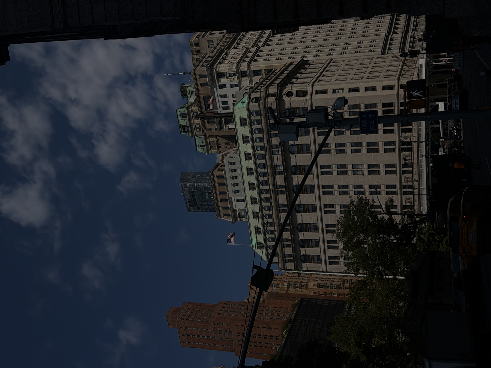 The image shows a street scene in New York City, United States of America, featuring several large buildings and a street with traffic.
In the foreground, on the right side of the image, a portion of a dark-colored building is visible. A traffic light with a pedestrian signal displaying a walking figure is prominent in the center of the frame. A blue sign with white lettering reading "PIER A" and an arrow pointing upwards is attached to the same pole as the traffic light.
Behind the traffic signal, a large, light-colored building with many windows dominates the mid-ground. It has a classical architectural style with decorative elements. To the left of this building, an American flag is visible flying from a pole on a green rooftop. Further to the left, a tall, reddish-brown brick building with numerous windows stands out. Smaller buildings and trees are interspersed between these structures.
The sky above is dark blue with scattered white clouds. The scene appears to be during the daytime, though the lighting suggests it might be late afternoon or early morning, or that the area is shaded. There are vehicles on the street, including a yellow taxi in the lower right portion of the image. Some people are visible on the sidewalk, but they are indistinct due to the resolution and lighting.