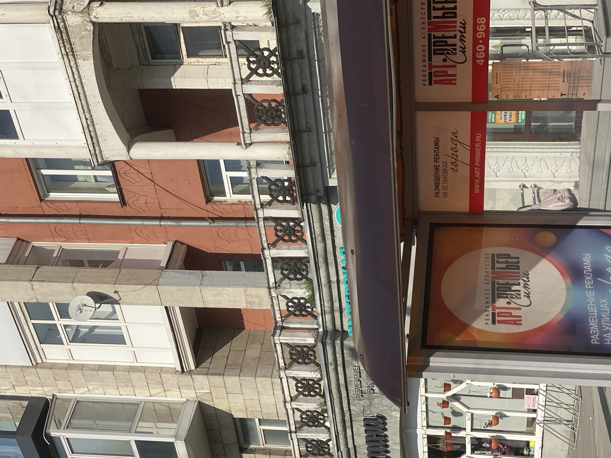 The image shows a bus stop shelter with advertisements on the side panels. Behind the shelter, there is a building with several floors and balconies. The building has a mix of architectural styles, with some parts appearing older and more ornate, while others are more modern. The advertisements on the bus stop shelter are for a company called "ART-PREMIER" which offers advertising services. The bus stop is located in Novokuznetsk, Russia, as indicated by the text on the advertisements. The scene appears to be during the daytime, with sunlight casting shadows on the ground. There are no people visible in the image.