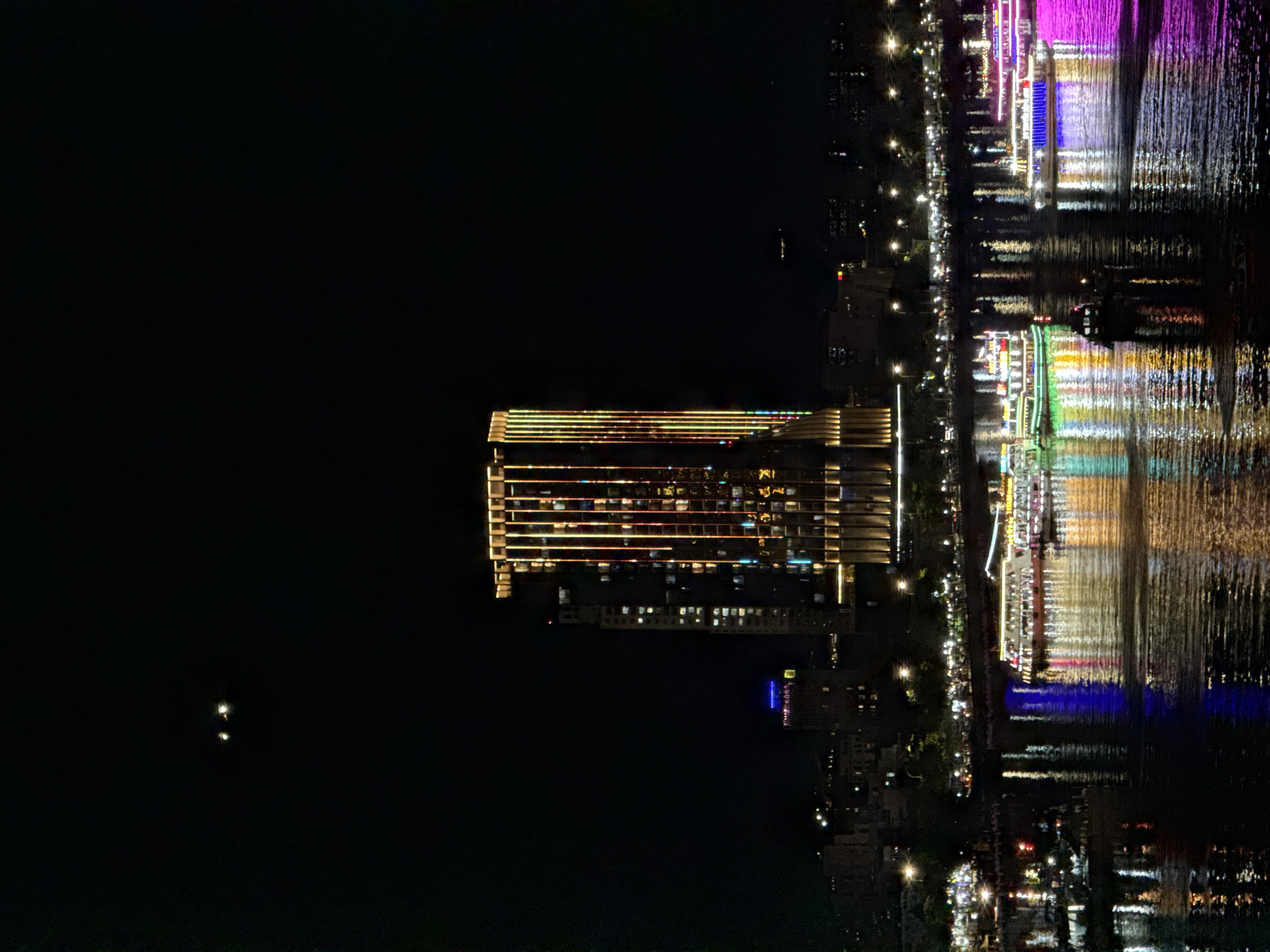 The image depicts a nighttime view of the Han River in Da Nang, Vietnam, with the Dragon Bridge and surrounding cityscape illuminated. In the foreground, several brightly lit tour boats navigate the river, their vibrant lights reflecting on the water's surface. These boats are a mix of styles and colors, with some featuring elaborate lighting displays.
Dominating the middle ground is a modern skyscraper, illuminated with vertical strips of colored lights that create a striking visual effect against the dark sky. To the left of this building, another, slightly shorter building is visible, with lights indicating windows and some blue lighting at its peak. Further in the background, the cityscape is a silhouette of buildings and lights, characteristic of a bustling urban environment at night.
In the upper part of the frame, against the black expanse of the night sky, two small, bright lights are visible, possibly distant aircraft or celestial objects. The overall scene is one of urban vibrancy and nighttime activity, showcasing the beauty of Da Nang's riverside architecture and illuminated infrastructure. The water reflects the lights of the city and the boats, creating a dazzling, colorful tableau.