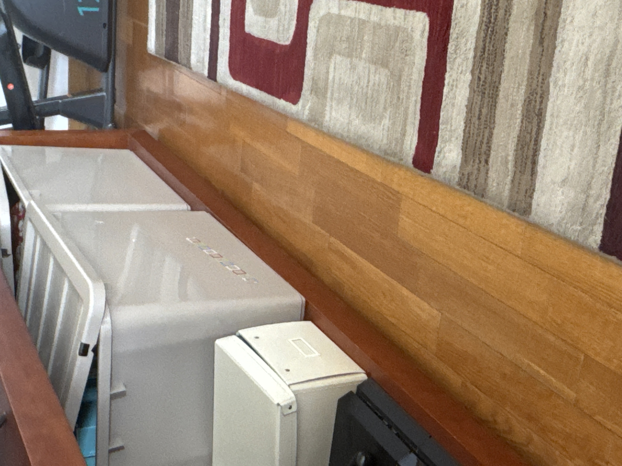 The image shows two light gray, rectangular storage boxes, likely plastic, with hinged lids. The box in the foreground is slightly open. The boxes sit on a surface, possibly the floor, adjacent to a wooden baseboard with a reddish-brown trim. Above the baseboard is a patterned rug or carpet in shades of white, beige, brown, and red. A black object is partially visible at the right edge of the image. Part of an exercise machine with a dark frame is seen in the upper left. The location in Mirandela, Portugal is unknown based solely on the visual information present.
