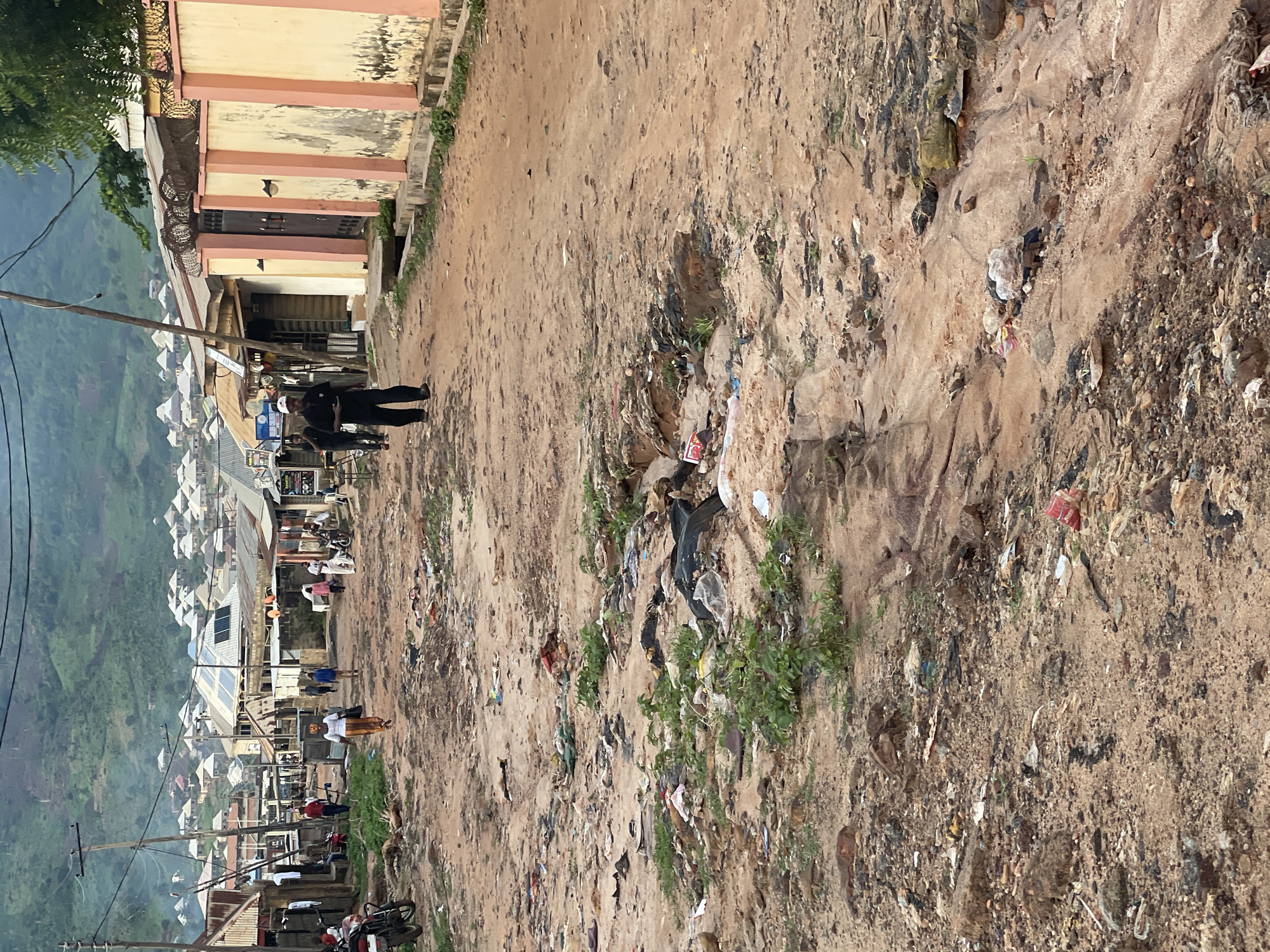 This image depicts a scene on a dusty, unpaved road in Lokoja, Nigeria, likely in a residential or commercial area on the outskirts of the city. The road is heavily littered with various types of trash, including plastic bags, bottles, and other debris, which has accumulated in a natural gully or erosion channel running through the scene. The surrounding environment shows a mix of structures, including low-rise buildings with corrugated metal roofs and some with solar panels, suggesting a developing urban area. There are also visible power lines and utility poles.
In the foreground, the focus is on the degraded state of the road and the surrounding land, characterized by dirt, rocks, and significant amounts of refuse. Several individuals are present in the midground and background, walking along the road. To the left, a person in a white top and orange skirt is walking away from the camera. Further down the road, a man in a black shirt and black pants, wearing a white cap, is walking towards the camera. Other individuals, some appearing to be walking in groups, are also visible in the distance.
The background reveals a more densely populated area with numerous houses stretching up a hillside, covered in greenery, indicating the hilly terrain of Lokoja. The overall impression is of a busy, albeit somewhat impoverished, community with challenges related to waste management and infrastructure.