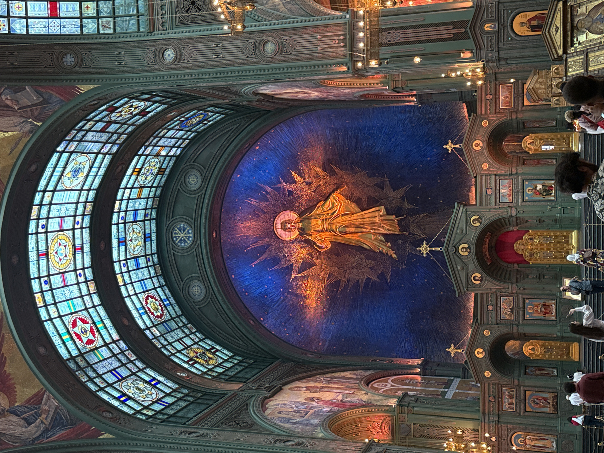 The image depicts the interior of the Main Cathedral of the Russian Armed Forces in Kubinka, Zelenograd, Russia. The grand scale of the cathedral is evident with its high, vaulted ceiling featuring stained-glass windows with intricate geometric and symbolic designs. Dominating the central wall is a large mosaic of Jesus Christ, rendered in gold and orange hues against a deep blue, star-studded background. The mosaic is framed by stylized, starburst-like elements.
Below the mosaic, a traditional iconostasis spans the width of the cathedral, featuring multiple tiers of icons housed within ornate, gilded structures. Arched doorways and decorative panels with religious imagery adorn the iconostasis. Several people are visible in the foreground, some appearing to be tourists taking photographs and observing the artwork. They are dressed in casual clothing. The overall lighting is dramatic, with spotlights illuminating the central mosaic and chandeliers casting a warm glow on the lower levels of the cathedral. The architectural style is distinctly Russian Orthodox, characterized by its opulence and religious symbolism.
