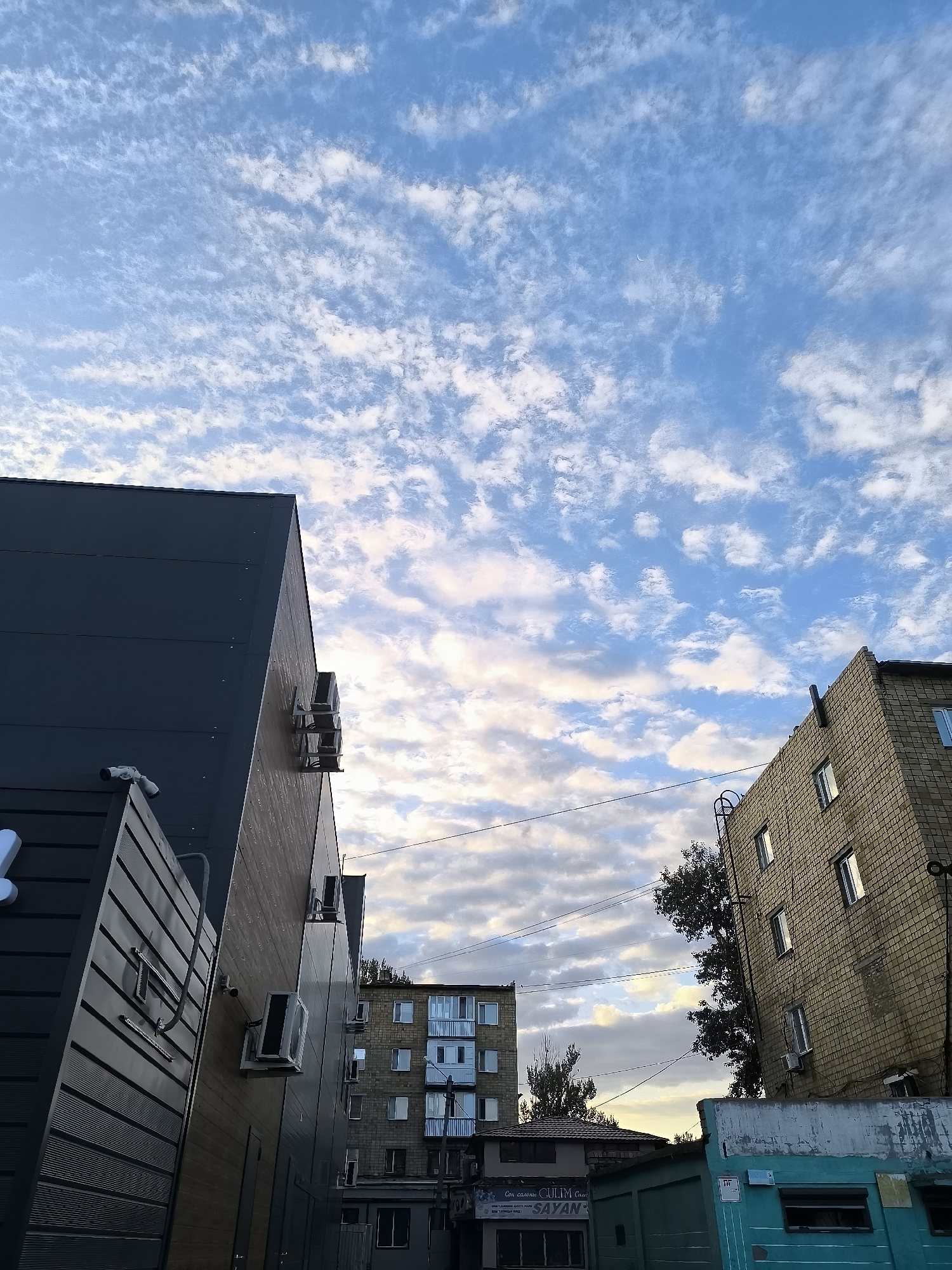 The image shows a view looking up at the sky with buildings in the foreground, set against a backdrop of a partly cloudy sky. On the left, a dark, modern building with a textured facade is visible, featuring several outdoor air conditioning units mounted on its side, as well as security cameras. Adjacent to it, another building with a similar dark, textured facade also has air conditioning units. Further into the background, a brick building with multiple windows and air conditioning units is seen, along with a more distant, multi-story residential building. Power lines crisscross the sky. A small crescent moon is visible in the upper portion of the sky. The scene appears to be in an urban environment.
