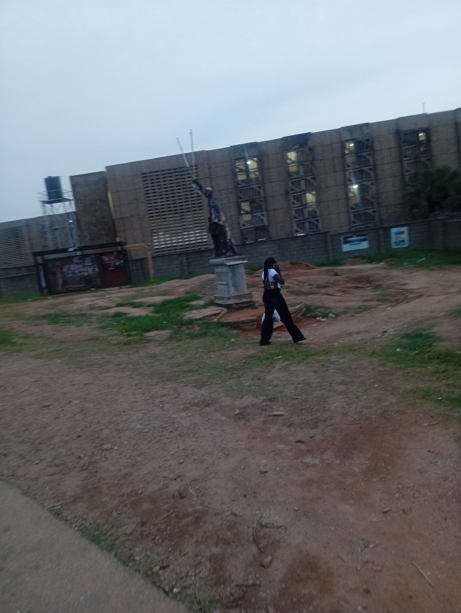 The image shows an outdoor scene with a building in the background and a statue in the middle ground. A person is walking in front of the statue. The building appears to be made of concrete and has many windows. A water tank is visible on top of a smaller structure to the left of the building. The ground in the foreground is dirt and grass. The sky is overcast. The scene is likely located in Jos, Nigeria, based on the style of the building and the overall environment.