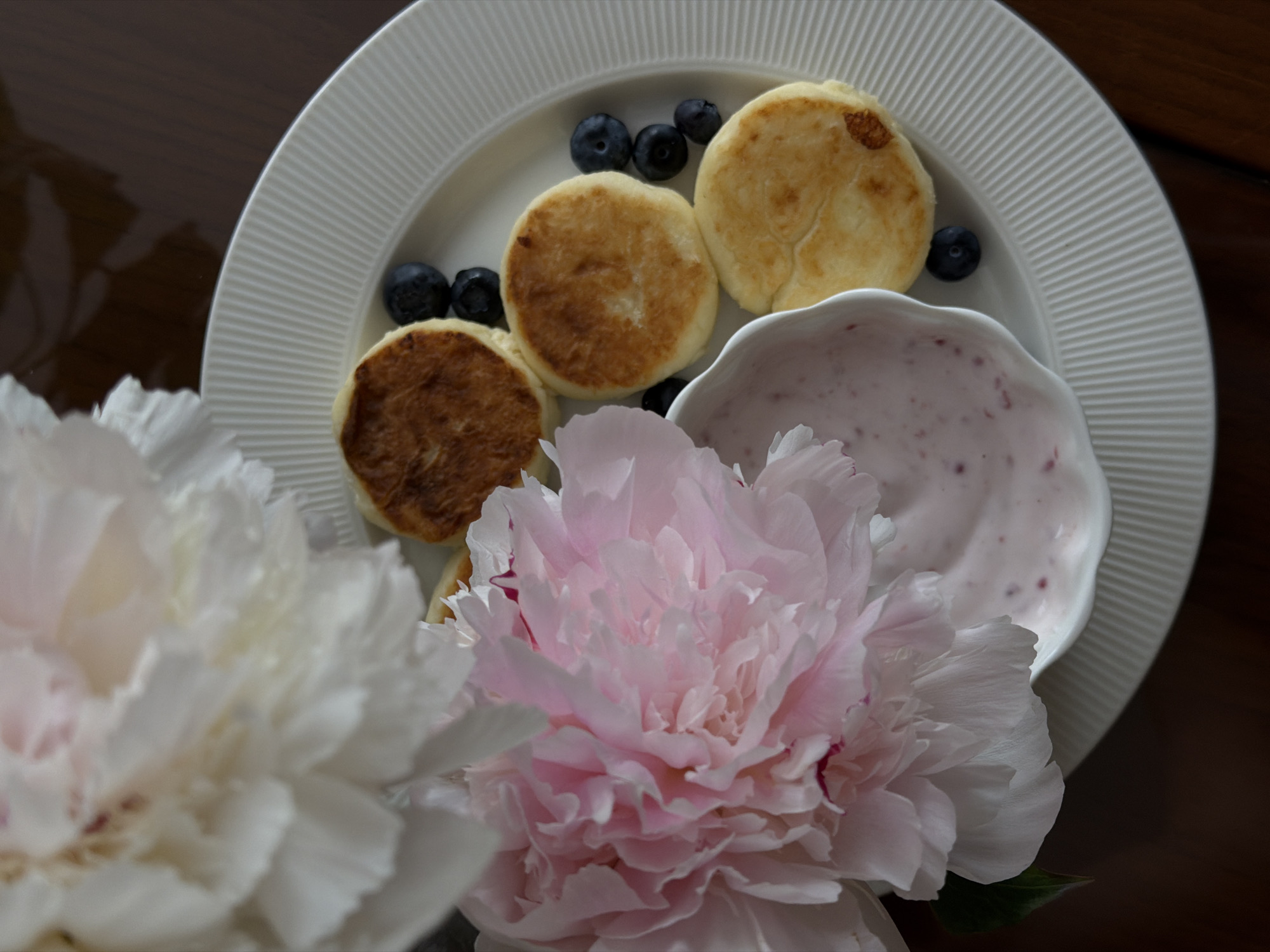 On a dark wooden surface, a white, ridged plate holds a still life composition. The plate contains four golden-brown pancakes, a small bowl of pink-speckled yogurt, and several blueberries. In the foreground, a white peony and a pink peony obscure a portion of the plate. The location is not identifiable as Budva, Montenegro based on the image.