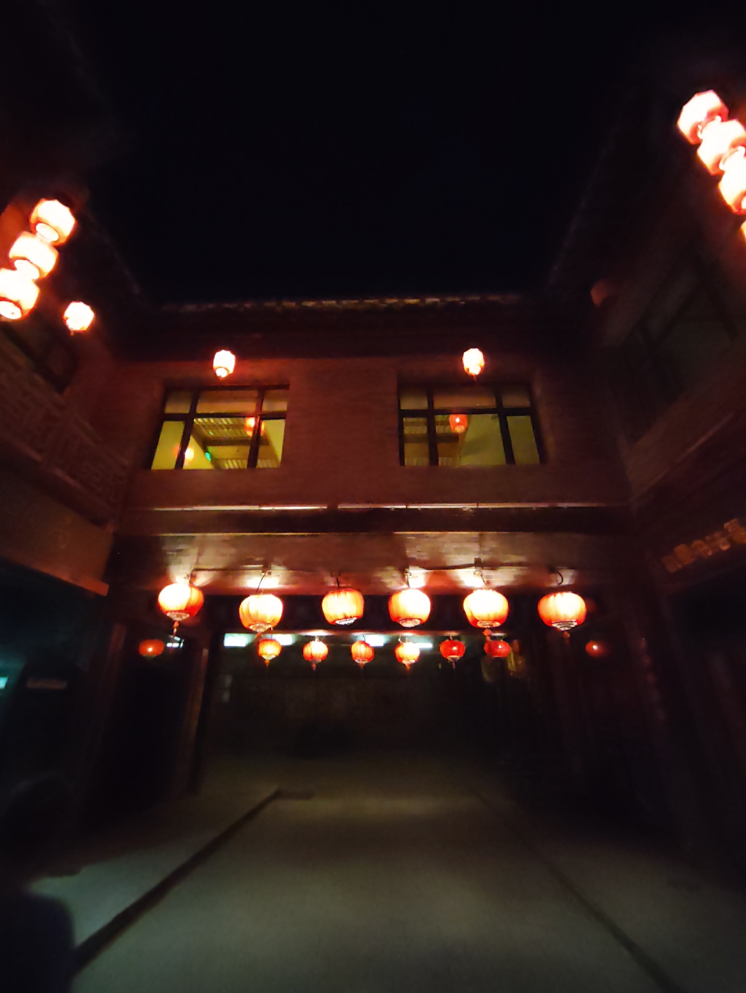 This is a night shot of a traditional street lined with buildings and decorated with numerous red lanterns. The lanterns are illuminated and hang in rows, casting a warm glow on the scene. The architecture appears to be of traditional Chinese design, with wooden elements and ornate details. Two windows are visible in one of the buildings, reflecting the light of the lanterns. The street itself is dimly lit, with a path leading into the distance. In the foreground, the silhouette of a person holding a long object is visible. The overall atmosphere is festive and traditional, evoking a sense of cultural heritage.