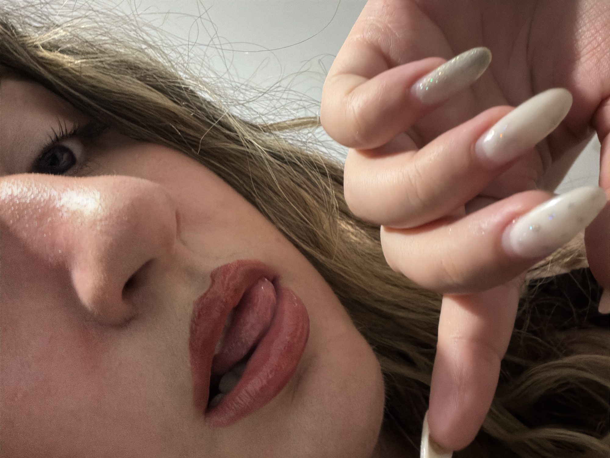 The image is a close-up, high-angle shot of a person's face and hand. The person has light skin, dark eyes, and blonde hair. Their mouth is open, with their tongue visible, and they are wearing lip gloss. Their fingernails are long and painted with a light, shimmery polish. The background is a plain, light-colored wall. There is no discernible location within the city of Plovdiv, Bulgaria, or any indication of it being in that city.