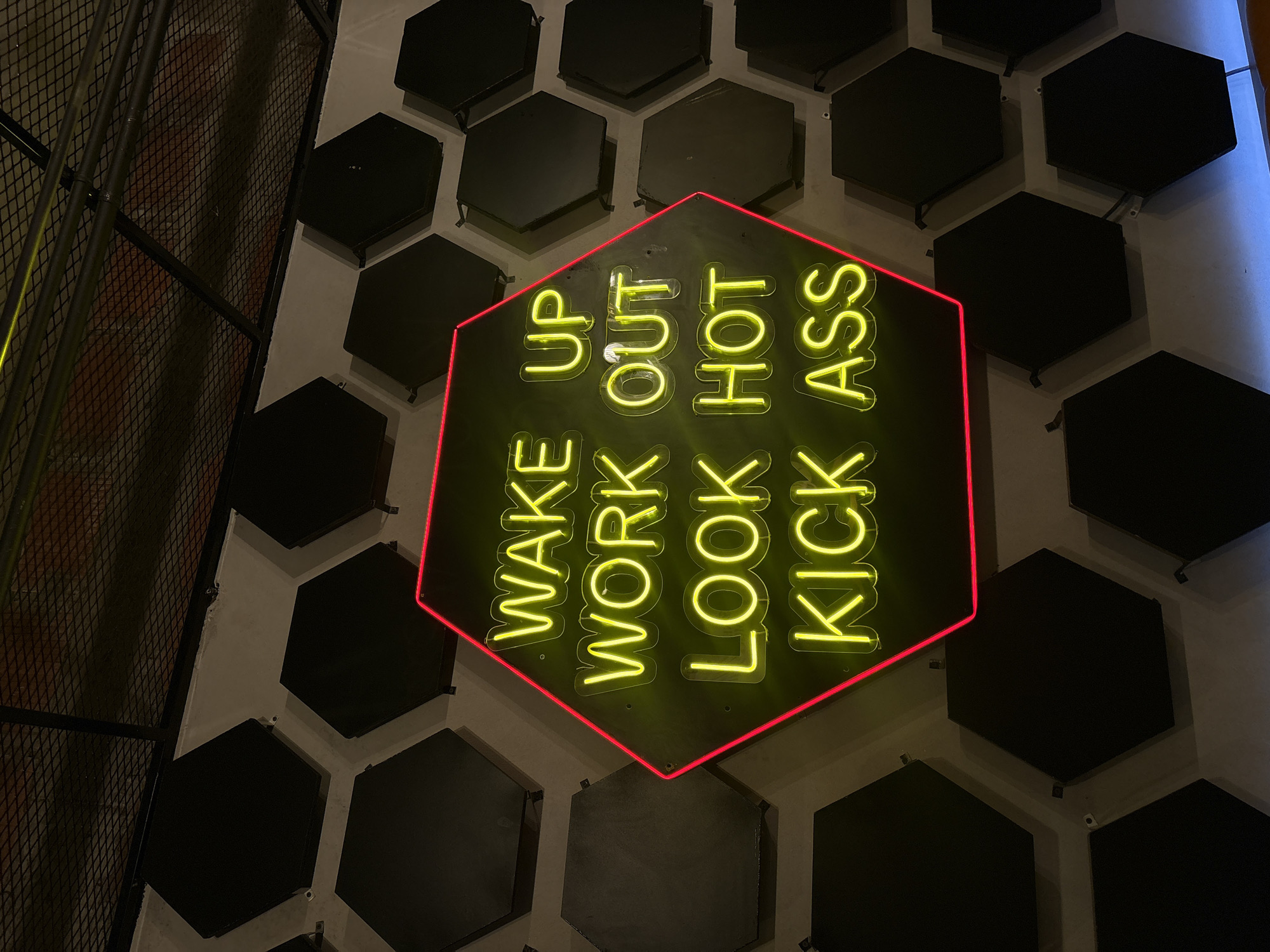 A medium, angled shot presents a wall adorned with hexagonal, black acoustic panels and a hexagonal neon sign with red trim, spelling out motivational phrases in stacked yellow text. The sign reads "WAKE UP," "WORK OUT," "LOOK HOT," and "KICK ASS." To the left, a black metal grate structure is partially visible. The location is inferred to be in San Bernardino Tlaxcalancingo, Mexico. There are no people present.