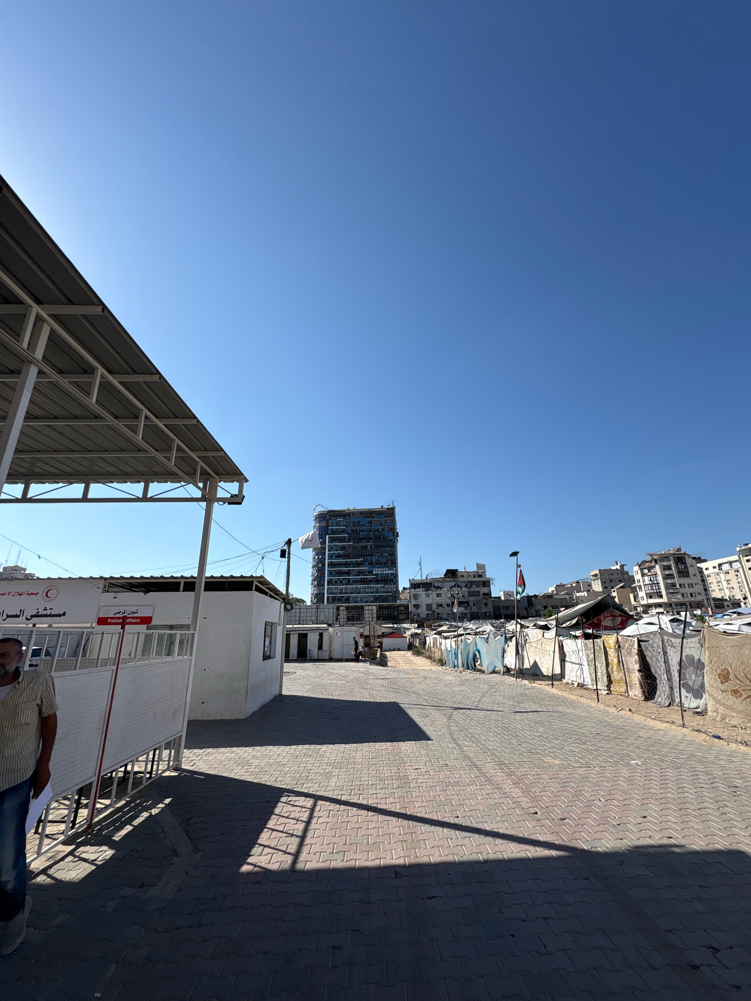 In the city of Gaza, Palestinian Territories, the scene depicts a street lined with makeshift tents and corrugated metal structures, suggesting a temporary or informal settlement. A paved road runs through the center, with shadows cast by an overhead canopy structure on the left side of the frame. A man in a striped shirt and jeans stands near a white fence with a red sign that reads "Patient Affairs." In the background, a modern high-rise building stands in stark contrast to the surrounding tents, some of which are adorned with fabric and blankets. A Palestinian flag is visible on a pole near the tall building. The sky is clear and blue, indicating a sunny day.