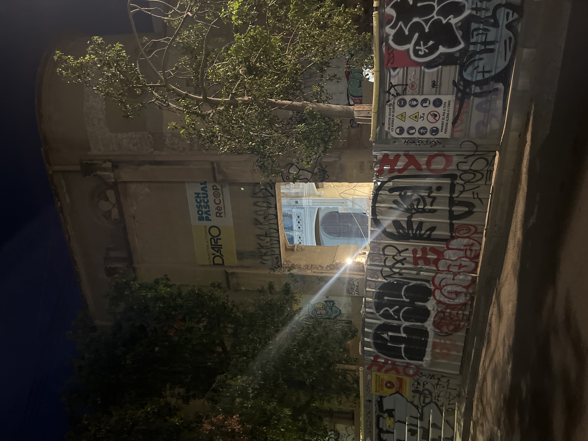 An image captured at night on a street in Barcelona, Spain, shows the facade of a building undergoing renovation. The building, partially obscured by trees, has a weathered appearance with some damage visible. A large banner advertising "BOSCH PASCUAL RECOP" hangs near the top of the facade. The lower portion of the building and the street are blocked by a metal fence covered in graffiti. The fence displays safety signs, including warnings about personal protective equipment and restrictions on unauthorized access. The street is dimly lit, with the main light source appearing to be a streetlamp casting a bright beam that creates a lens flare effect in the photograph. The overall scene depicts an urban environment with signs of construction, artistic expression through graffiti, and nocturnal lighting.