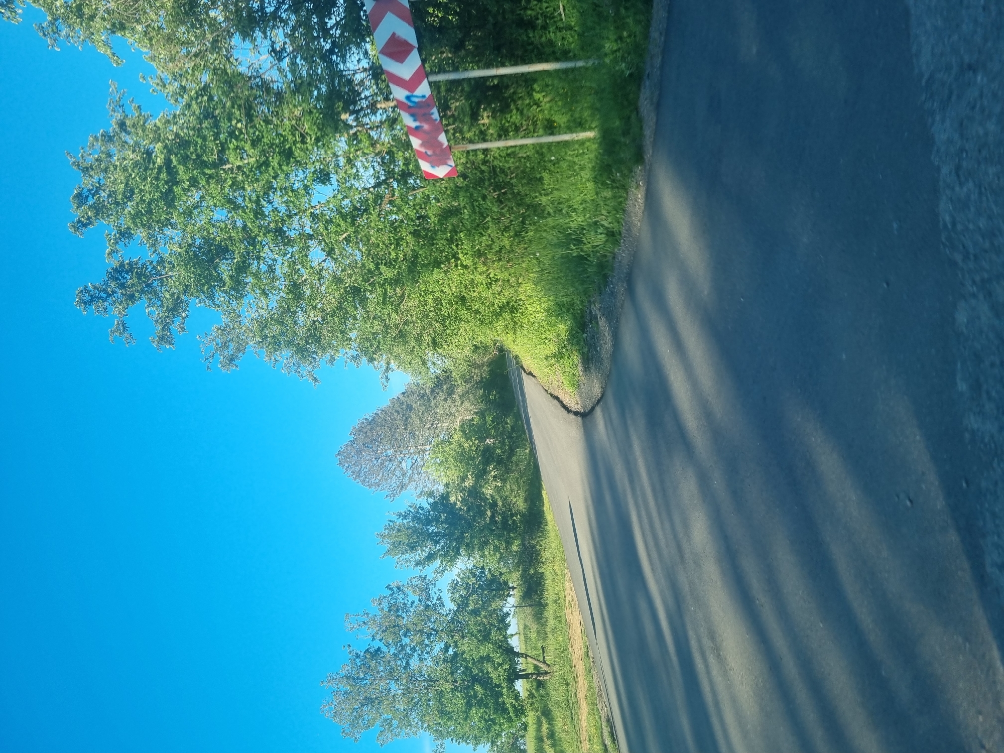 The image depicts a narrow asphalt road winding through a rural area. On the left side of the road, there is a clear blue sky partially obscured by leafy green trees. A red and white striped hazard marker stands near the roadside. Vegetation, including grass and bushes, lines the edge of the road. The road itself is cast in shadow from the trees. The location is Sheremetia, Bulgaria.