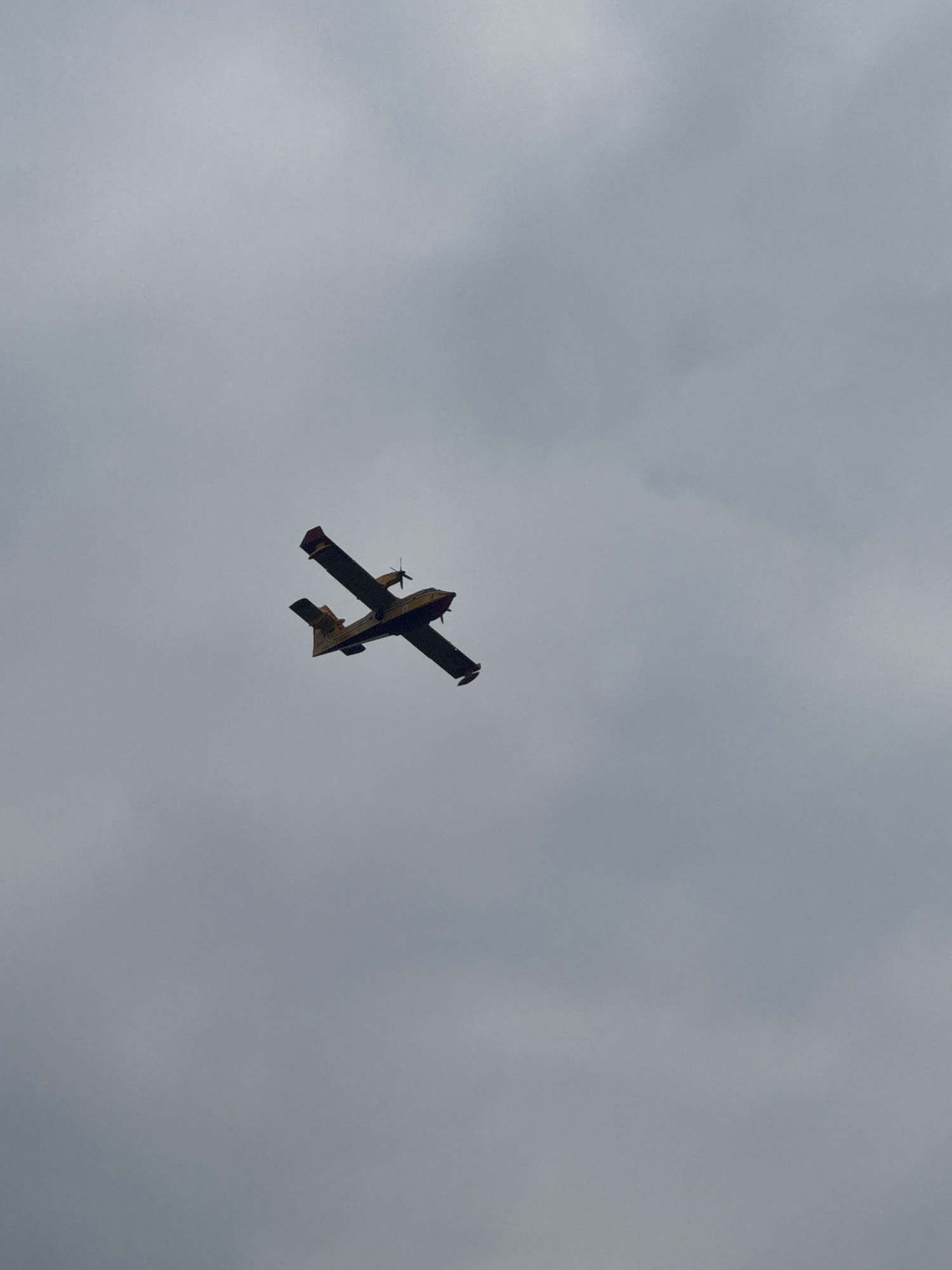 A low-angle, vertical shot captures a yellow and red amphibian aircraft flying against a heavily overcast, grey sky. The aircraft is positioned in the upper right quadrant of the frame, with its wings extended and propellers visible. The overcast sky suggests a potentially damp or stormy weather condition over the city of Como, Italy, though no specific landmarks of Como are visible in the frame. The scene is devoid of people and any other discernible objects. The overall impression is one of an isolated aircraft against a uniformly cloudy backdrop.