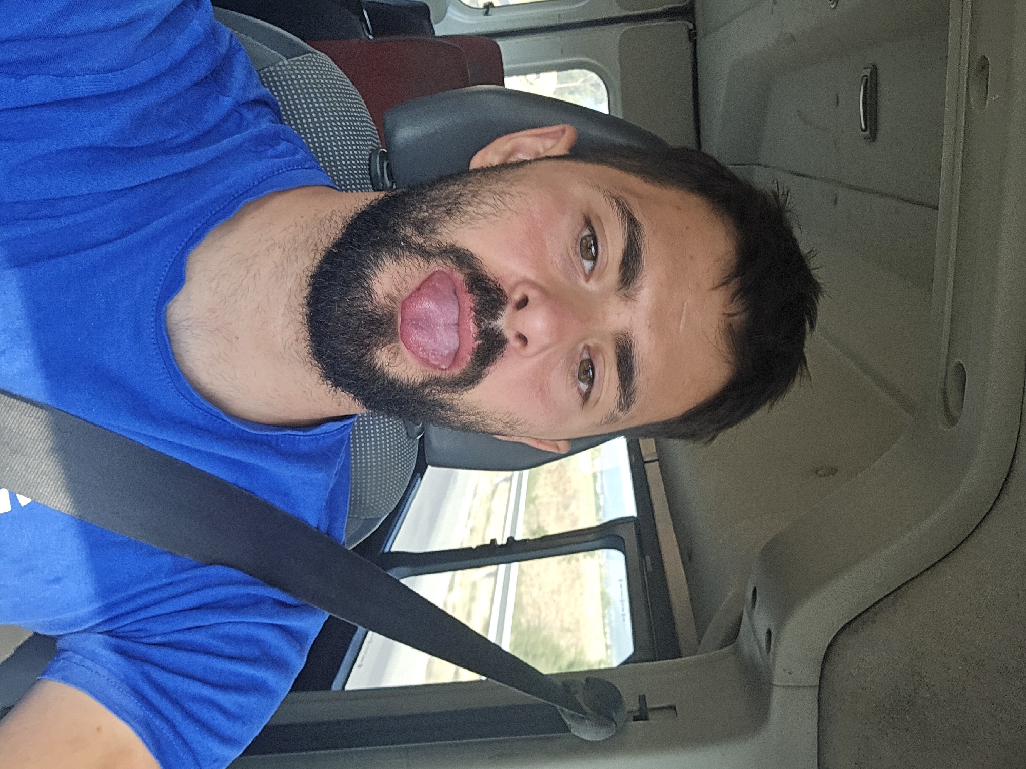 A man with a beard and dark hair sticks his tongue out while sitting in the passenger seat of a vehicle. He is wearing a blue t-shirt and a black seatbelt. The interior of the vehicle, including a headrest and a ceiling panel, is visible. Outside the window, a road and dry, grassy landscape can be seen. The location is identified as Konare, Bulgaria.