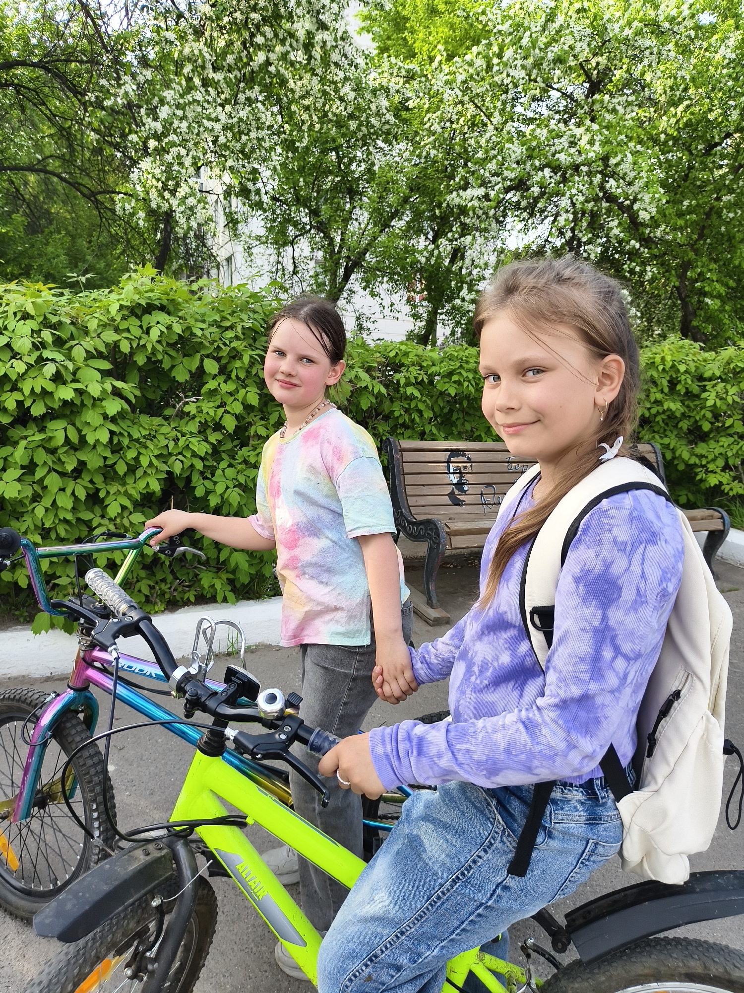 Two girls stand with bicycles on a paved surface, likely a sidewalk or path. The girl on the left wears a tie-dye shirt and gray pants, standing beside a bicycle with a multi-colored frame. The girl on the right is seated on another bicycle wearing a purple tie-dye long-sleeved shirt and jeans, with a white backpack slung over her shoulder. They are holding hands. Behind them are green bushes, trees with white blossoms, and a bench with a drawing on it. The scene appears to be outdoors in a city park or residential area.