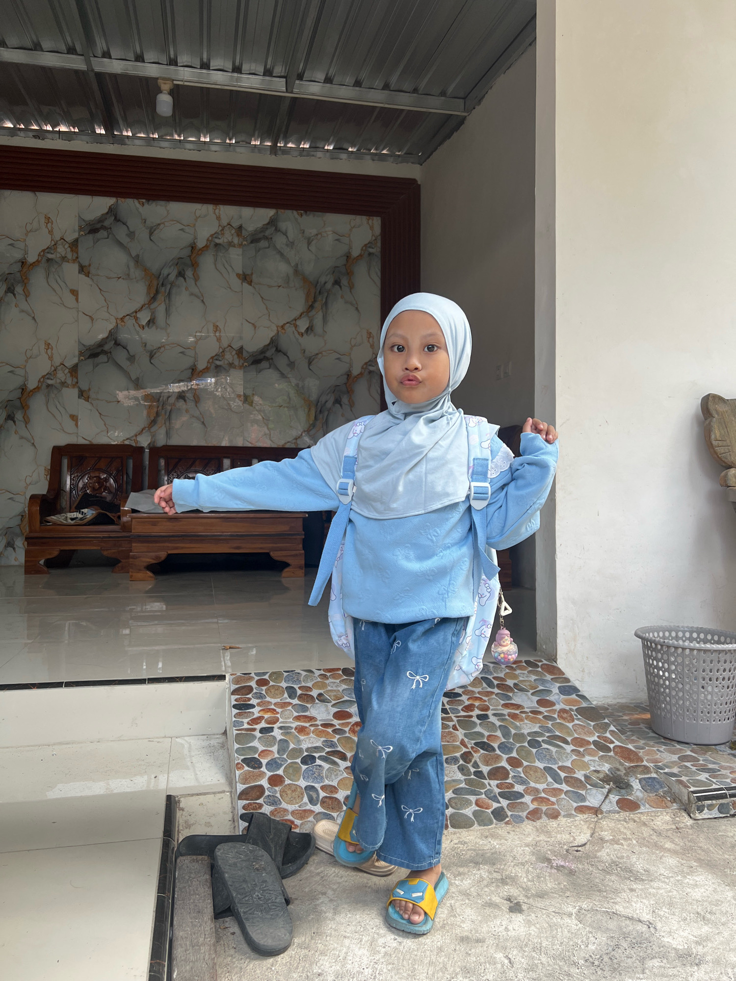 A young girl wearing a light blue hijab, matching sweatshirt, and blue jeans with a bow pattern stands outside. She has a light blue backpack with white straps and a charm attached to it. The girl is standing on a mosaic-tiled step, with a concrete ground and a building in the background. To the left of the girl, there are wooden chairs and a table with patterned marble walls behind them. To the right, a grey plastic waste bin is visible. The scene appears to be in front of a residential building in Kecamatan Pati, Indonesia, given the architectural style and the presence of what looks like an outdoor veranda.