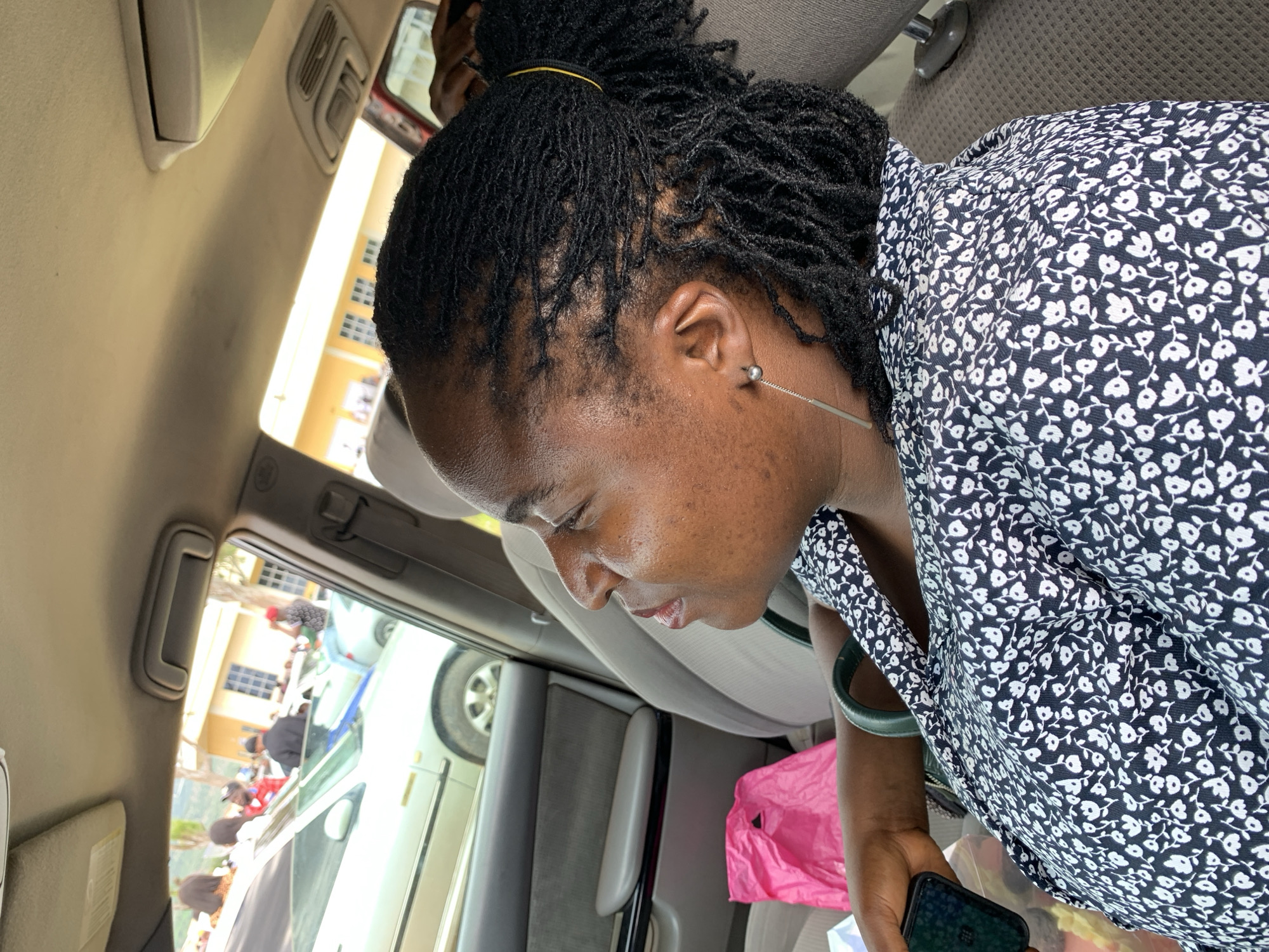A medium shot captures a woman from the chest up, seated inside a car, looking out the window. She has dark skin and braided hair pulled into a ponytail, adorned with a yellow hair tie. Her attire consists of a dark blue shirt with a white floral pattern. She is wearing a long, silver earring. In her left hand, she holds a black smartphone. To her left, a pink plastic bag rests on the car seat. The background shows other cars and buildings, suggesting an outdoor urban setting, likely in Yelwa, Nigeria. The overall scene is well-lit, with the interior of the car visible, including the car's upholstery and part of the dashboard.