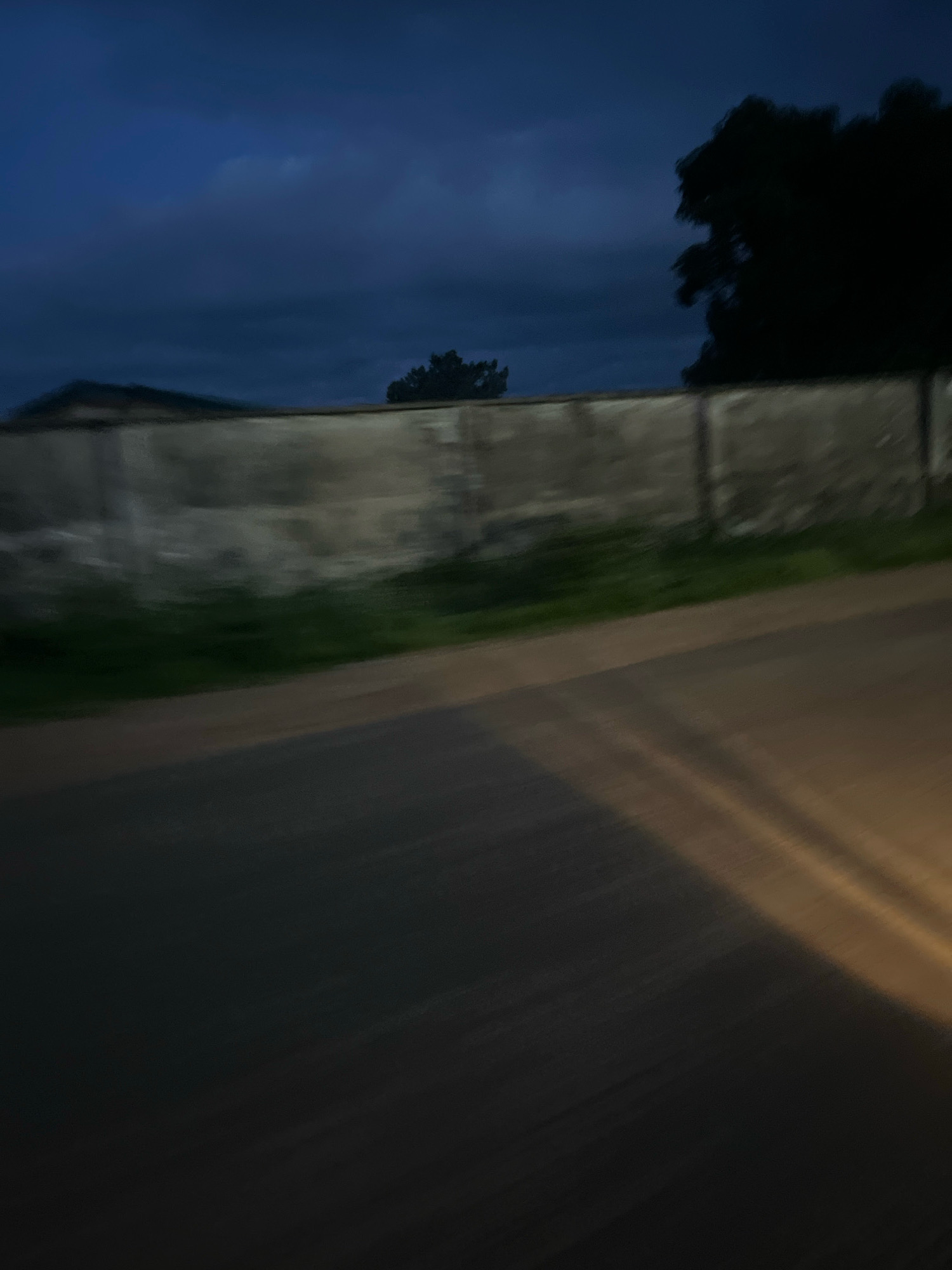 The image depicts a low-light, motion-blurred view of a street scene in Jos, Nigeria, likely taken at dusk or early dawn. A weathered, concrete wall runs horizontally across the middle ground, with some vegetation visible behind it. Silhouettes of trees are present against a dark blue sky with some clouds. The foreground shows a portion of a road, with a streak of light, possibly from a headlight, diagonally across it. The overall impression is one of movement and low visibility.
