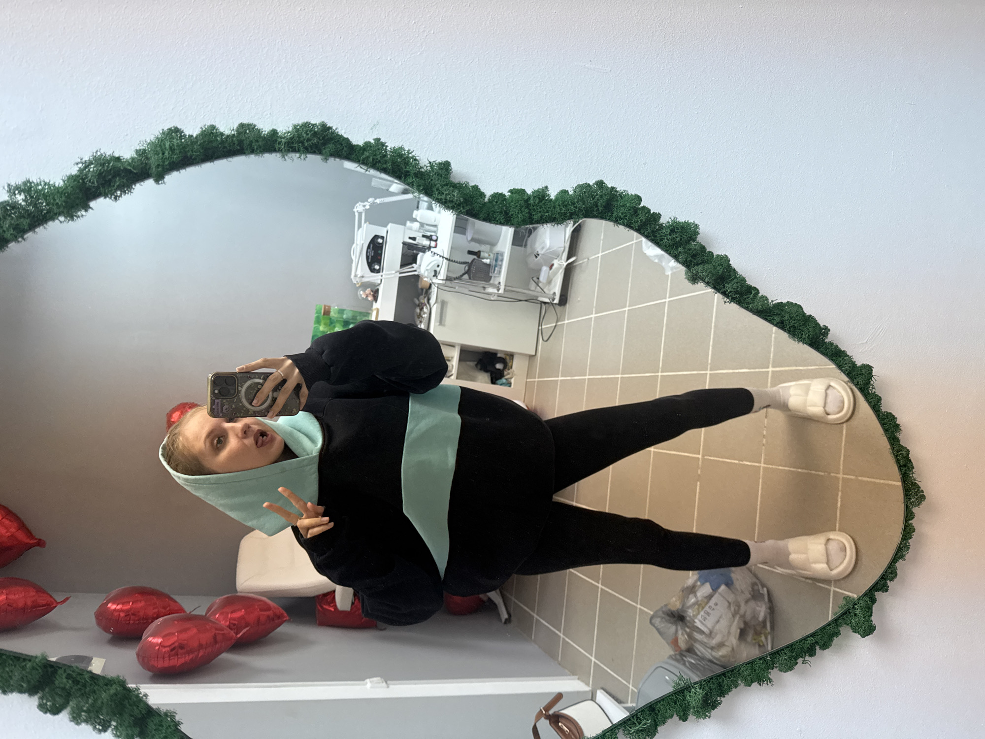 The image captures a playful mirror selfie of a young woman, rotated 90 degrees counter-clockwise. The main subject is a young woman, likely in her late teens or early twenties, with blonde hair, looking directly into the camera with a cheerful, slightly mischievous expression, her tongue sticking out. She is dressed in a black hoodie featuring a light blue horizontal stripe and a matching light blue inner hood, paired with black leggings, white socks, and cream-colored slide sandals or slippers. She holds a smartphone with a patterned case in her right hand to take the photo, while her left hand forms a peace sign.
The selfie is taken in a distinctive heart-shaped mirror, which is bordered by a lush green moss or artificial foliage. The reflected setting is an indoor space with light grey or beige tiled flooring and light-colored walls. Scattered on the floor to the left of the woman are several shiny red heart-shaped balloons. In the background reflection, what appears to be a counter or workstation holds various white electronic devices or appliances, suggesting a commercial environment such as a studio, salon, or office. A white plastic chair and a clear plastic bag containing items are also visible within the reflection. The time of day is not indicated, and the overall atmosphere is casual and lighthearted. The location context for this scene is Izhevsk, Russia. No legible text is visible in the image.