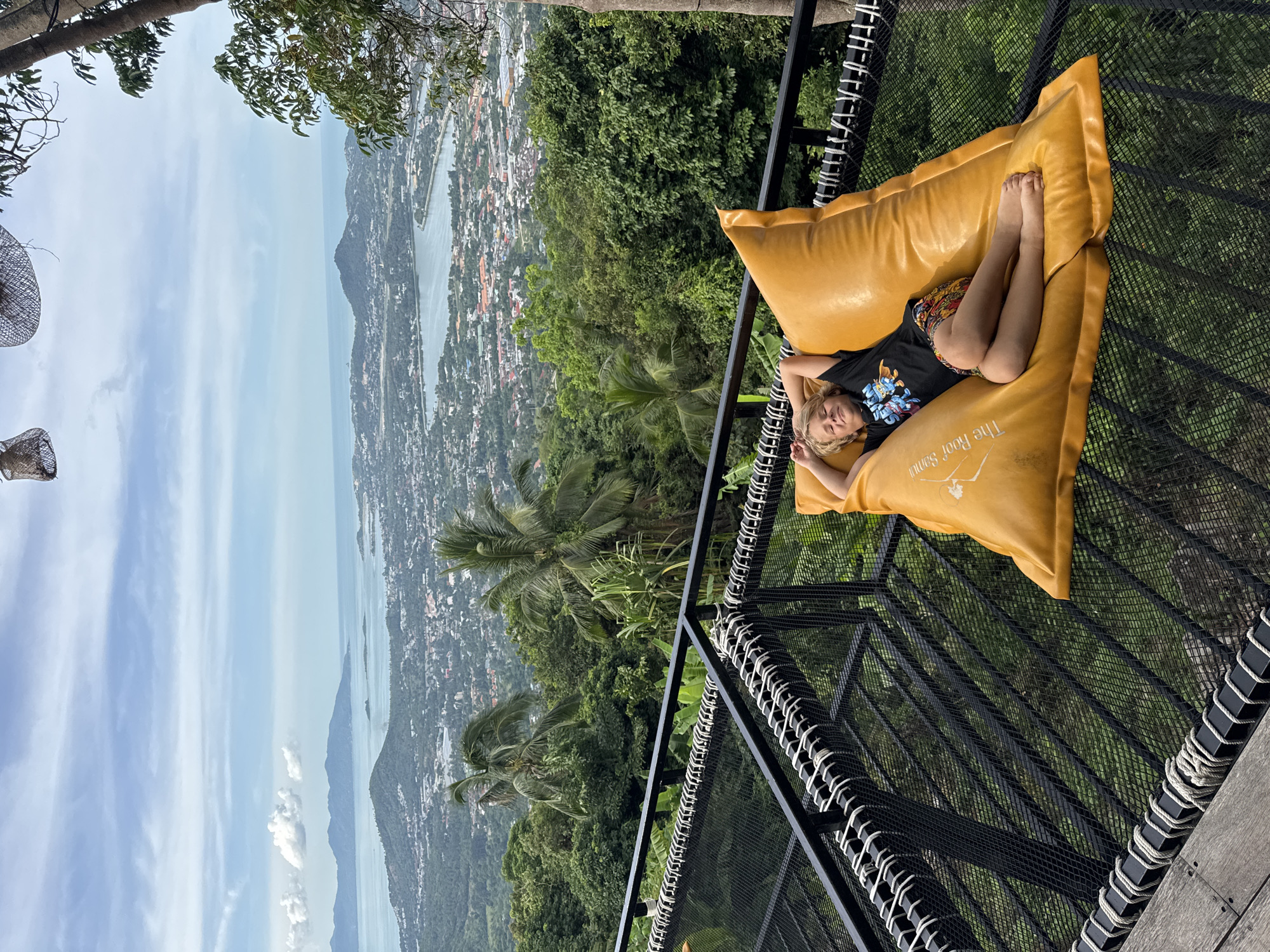 In an outdoor setting with a panoramic view of a city and ocean, a young person is relaxing on a large, mustard-yellow beanbag. The beanbag is situated on a black netted platform, possibly a balcony or observation deck, which is bordered by a black metal railing. The person is lying on their back with their head resting on their hands and their eyes closed, appearing to be asleep or resting comfortably. They are wearing a black t-shirt with a graphic design and patterned shorts. The background reveals a densely vegetated hillside with palm trees, leading down to a city with buildings and a body of water that meets the ocean. The sky is partly cloudy with a pale blue hue. The scene suggests a leisurely moment in a scenic location.